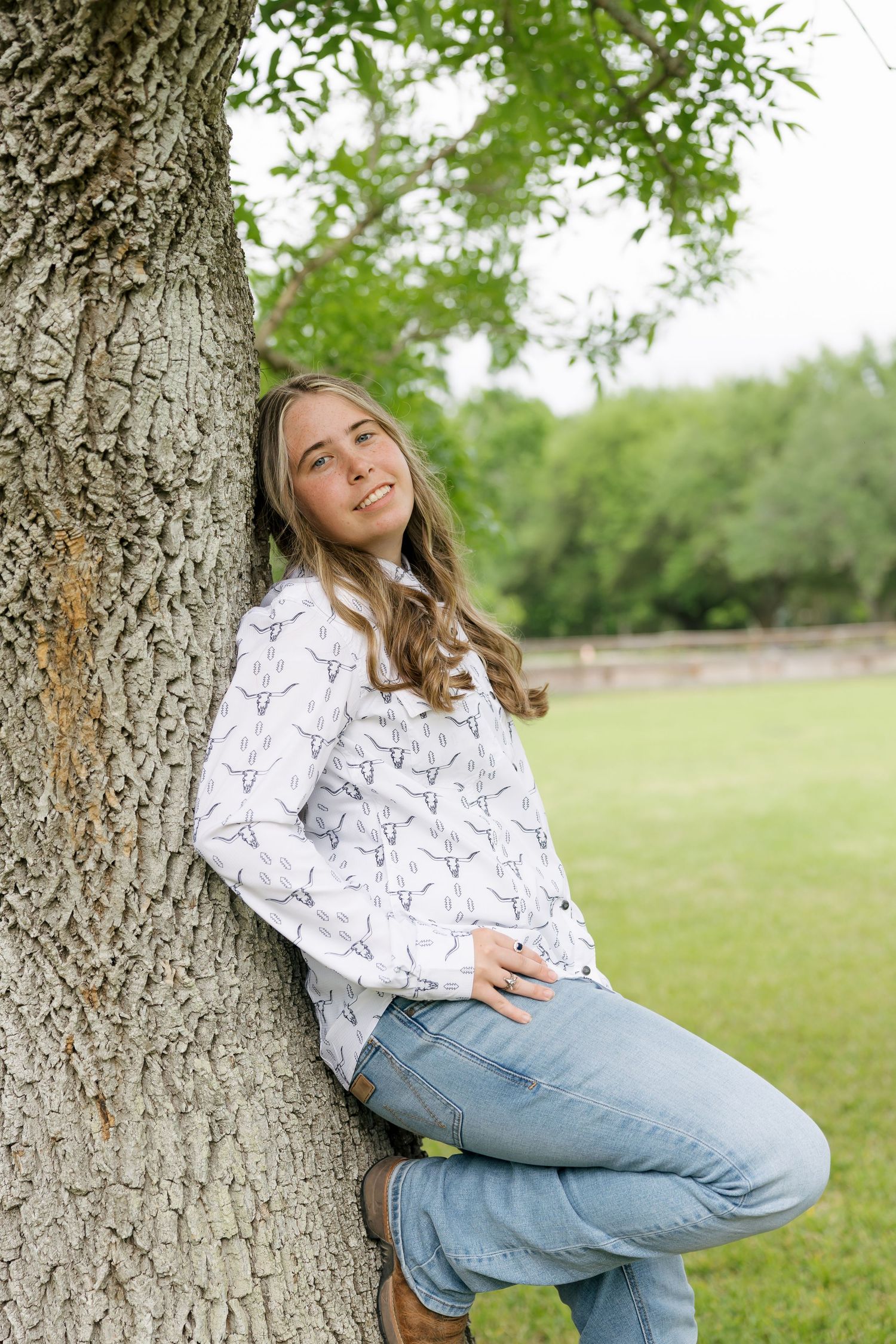 Young woman in patterned shirt and jeans leaning against a tree in a green park setting.