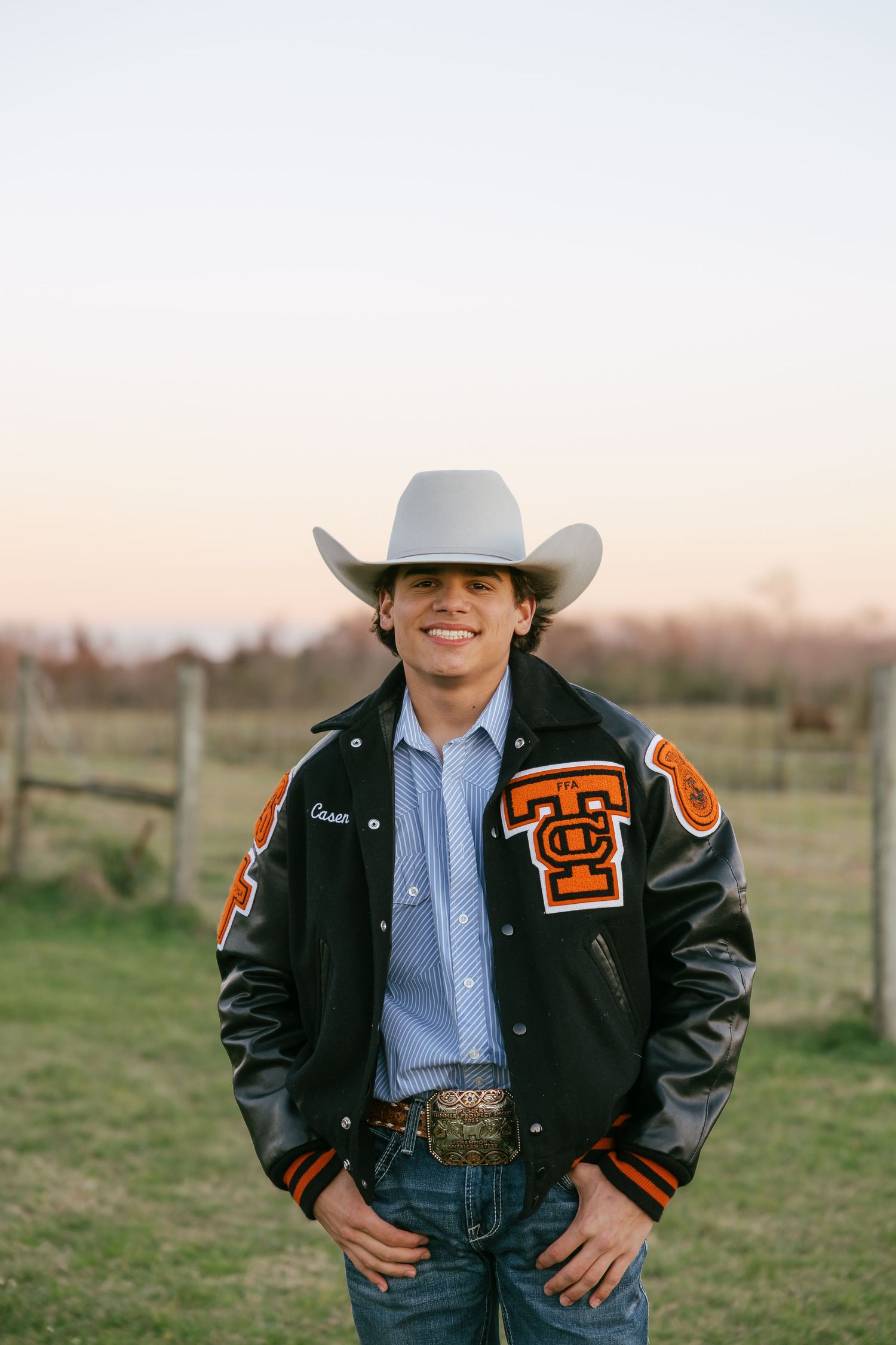 Smiling young man in cowboy hat and varsity letterman jacket posing at golden hour on a ranch.