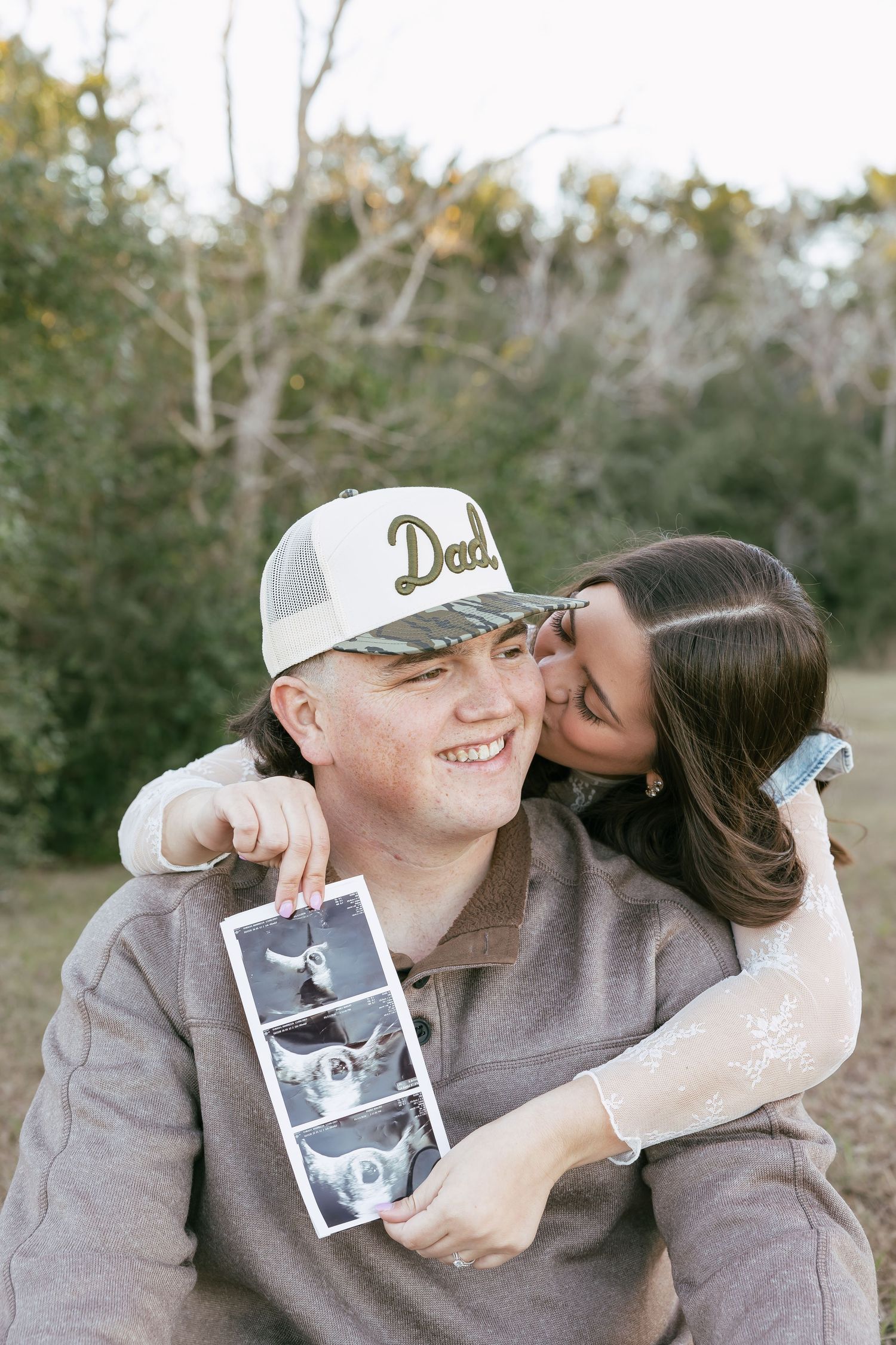 A smiling couple shares a pregnancy announcement outdoors, the man wearing a Dad hat and holding ultrasound photos.
