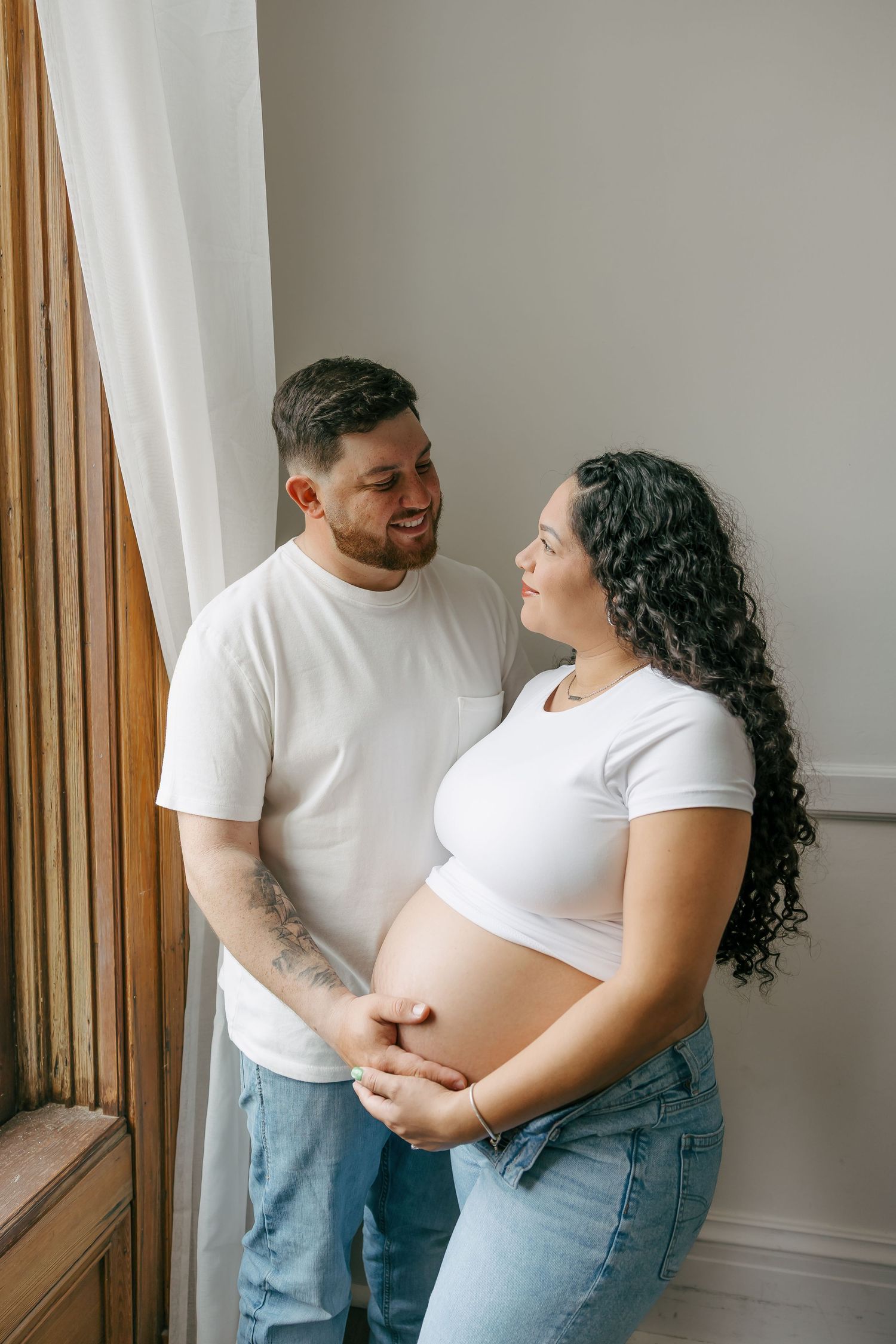 A smiling pregnant couple in matching white shirts holds her baby bump while standing together near a curtained window.