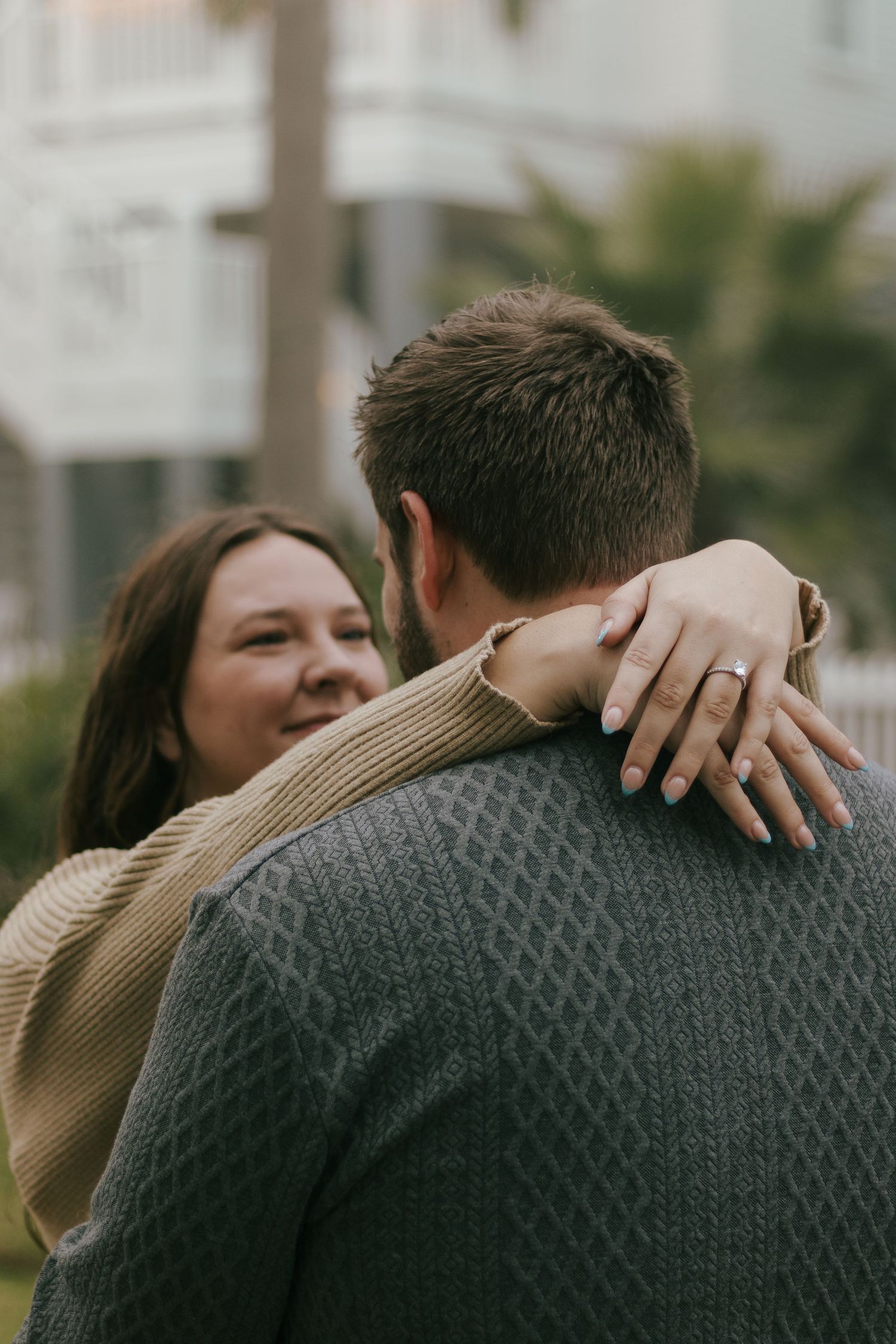 A woman with an engagement ring smiles while embracing a man from behind near palm trees and a white building.