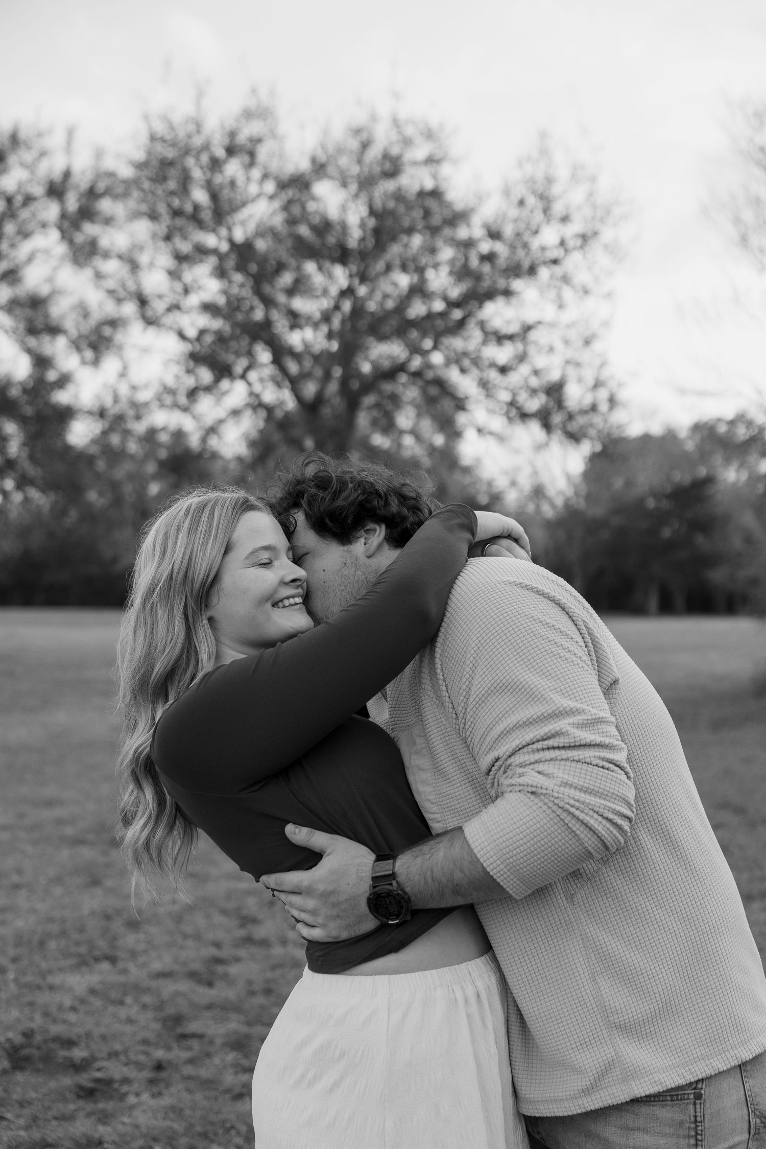 Black and white photo of laughing blonde woman being hugged by man in park with bare trees.