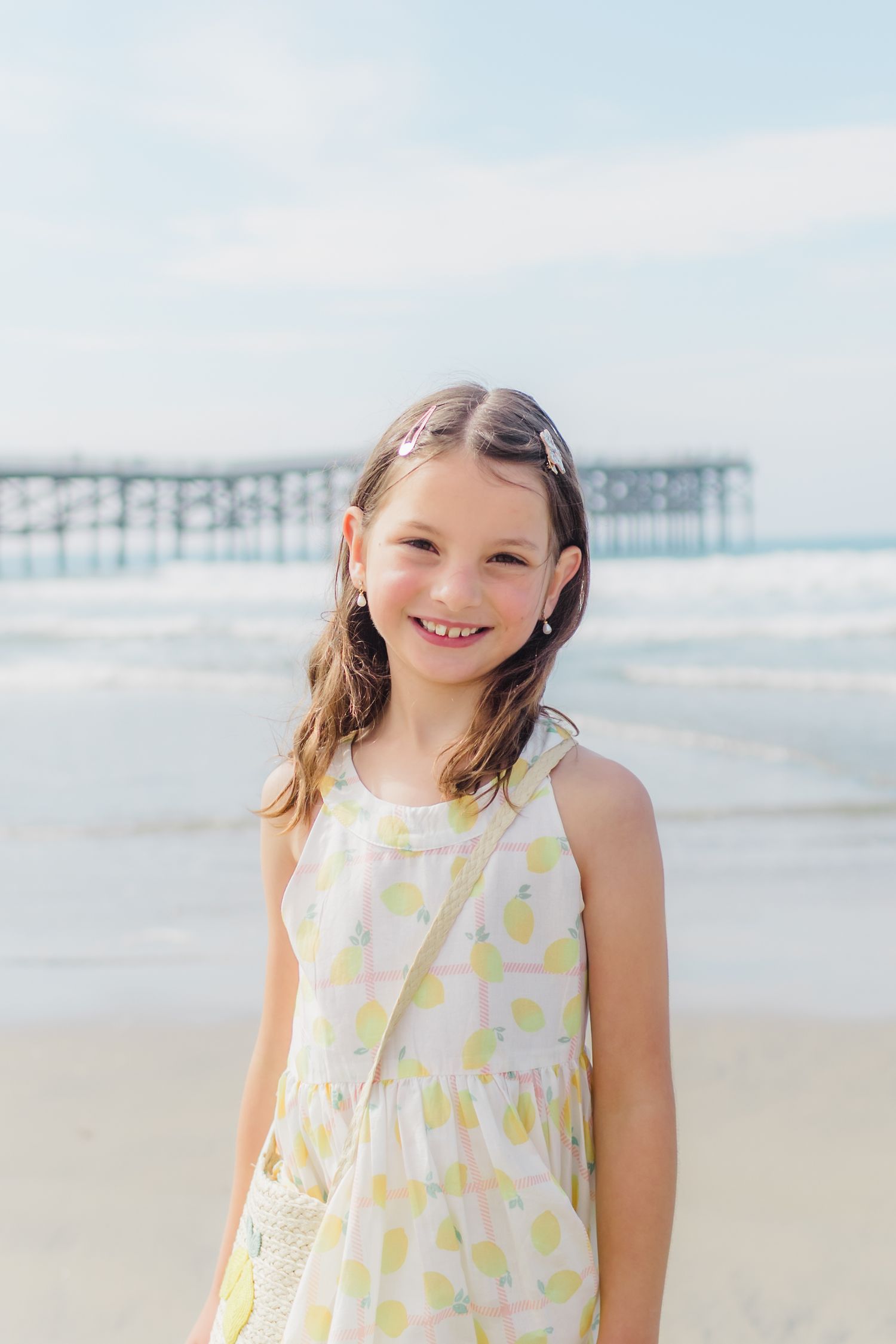 A child in a yellow and white summer dress stands on the beach with a pier visible in the background.