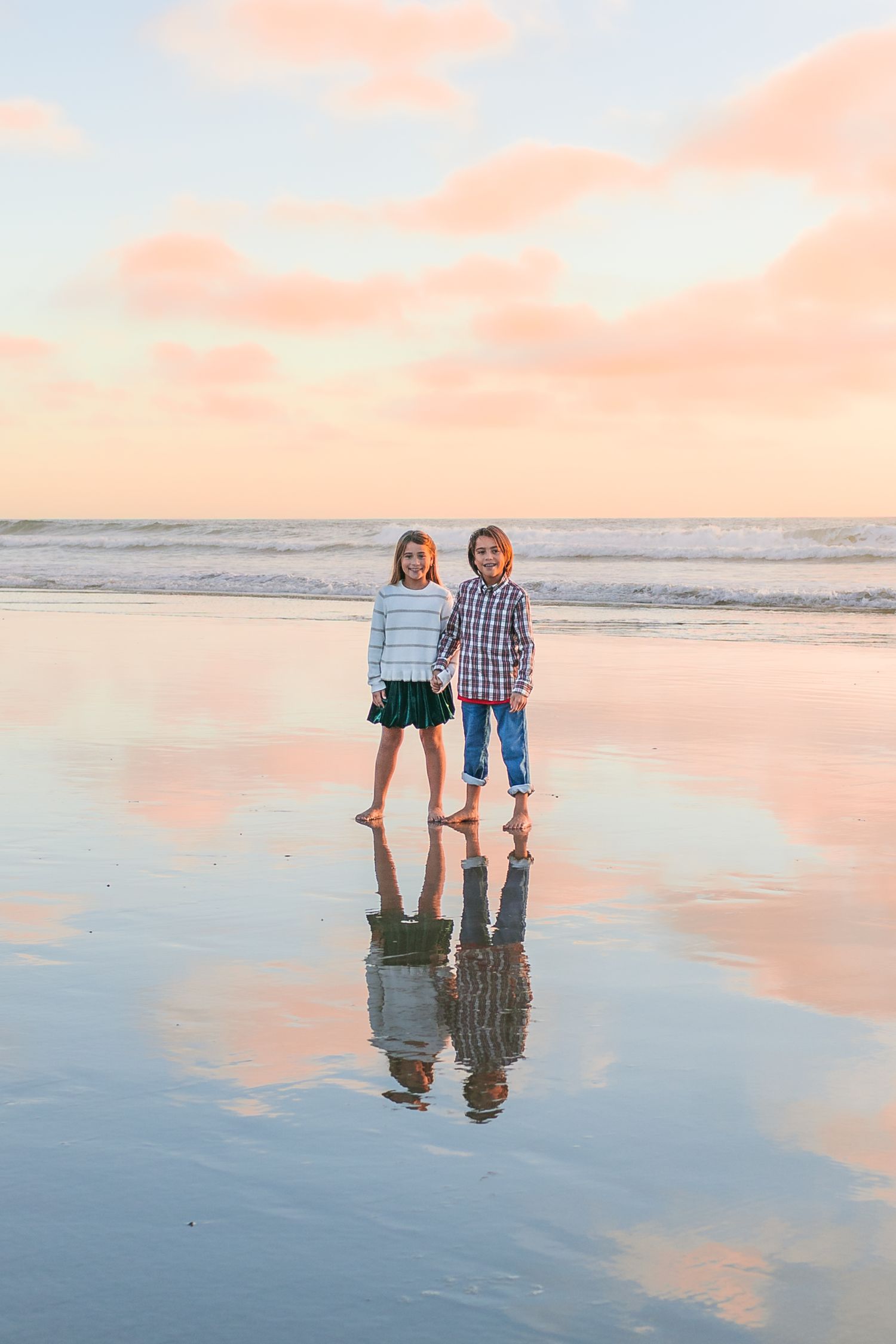 Two people walking along a reflective beach shoreline at sunset with pink and orange sky.