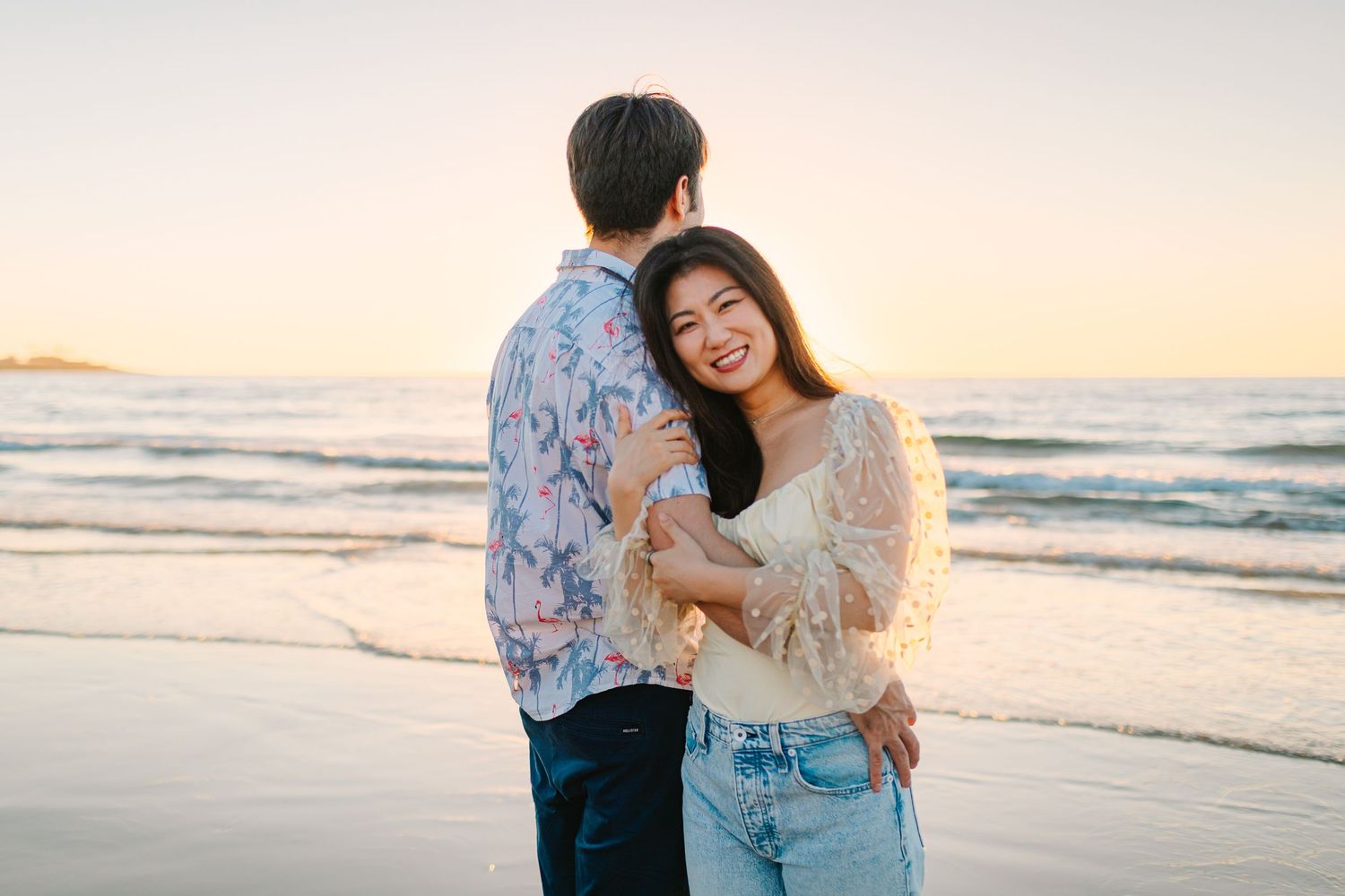 Couple embraces on sandy beach during golden sunset with ocean waves in background.
