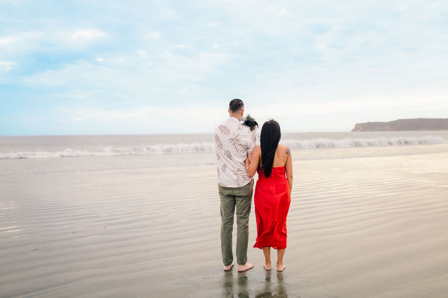 A couple walks together on a sandy beach with mountains in the distance during a cloudy day.