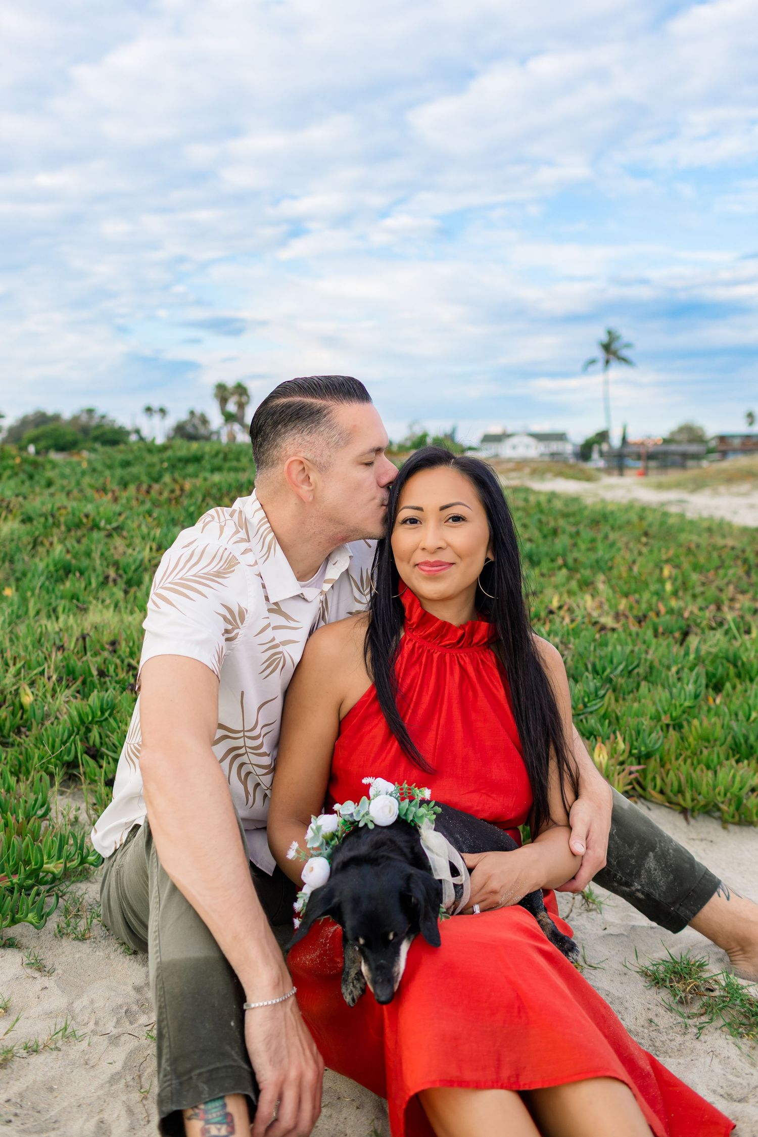A couple shares a romantic moment on the beach, with the woman wearing a bright red dress and holding a small plush toy.