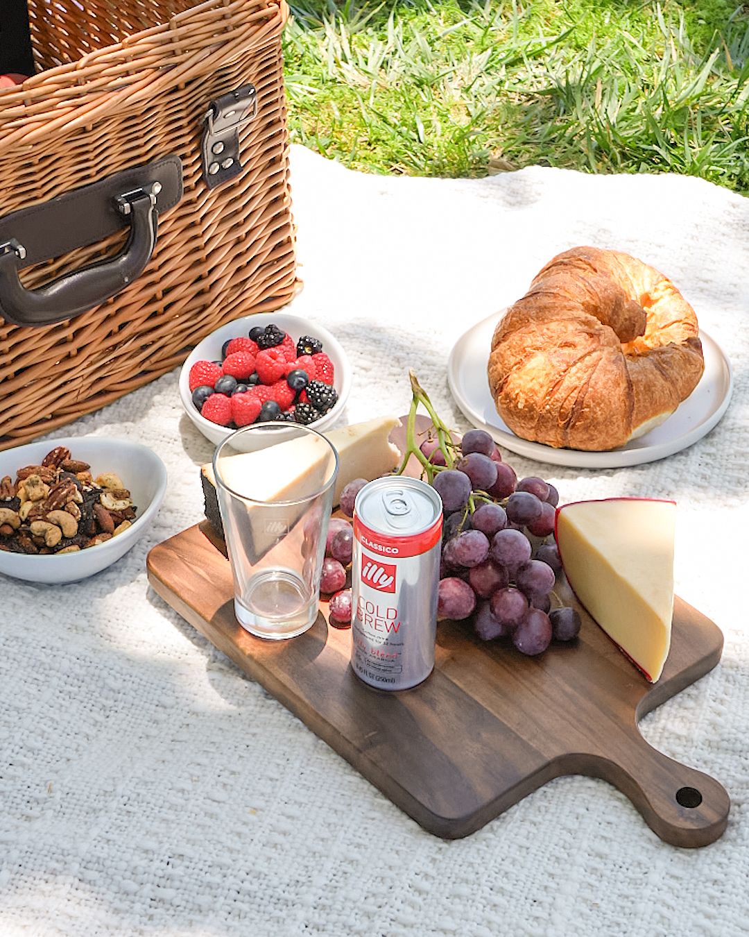 Picnic spread on white blanket with charcuterie board featuring berries, grapes, cheese, croissant and a Diet Coke can.