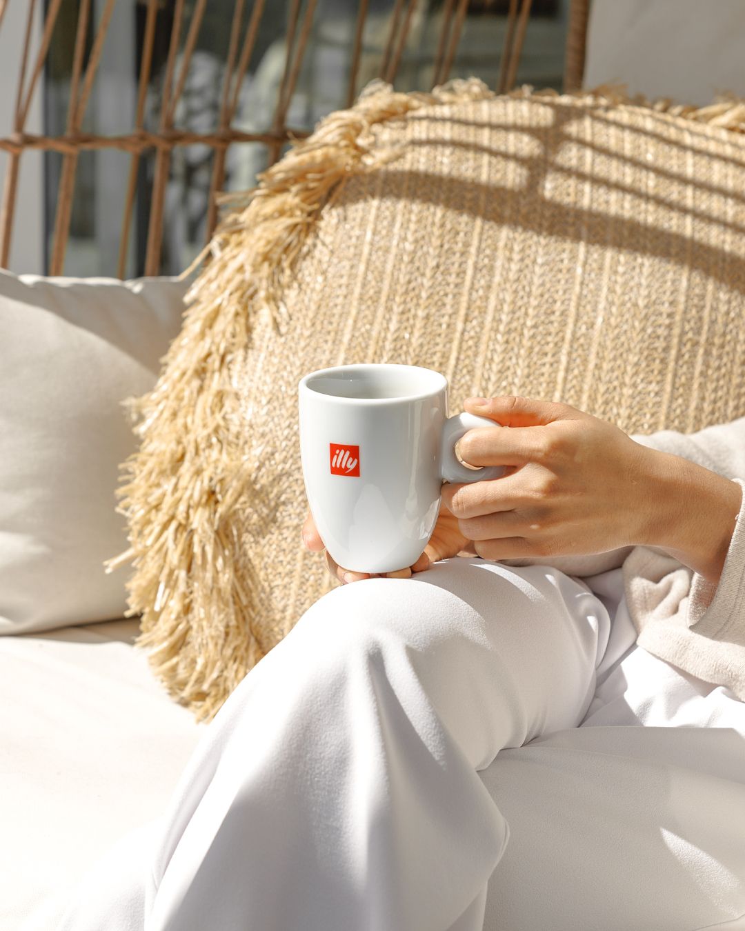Hand holding white illy coffee cup against beige natural decor and pampas grass background.