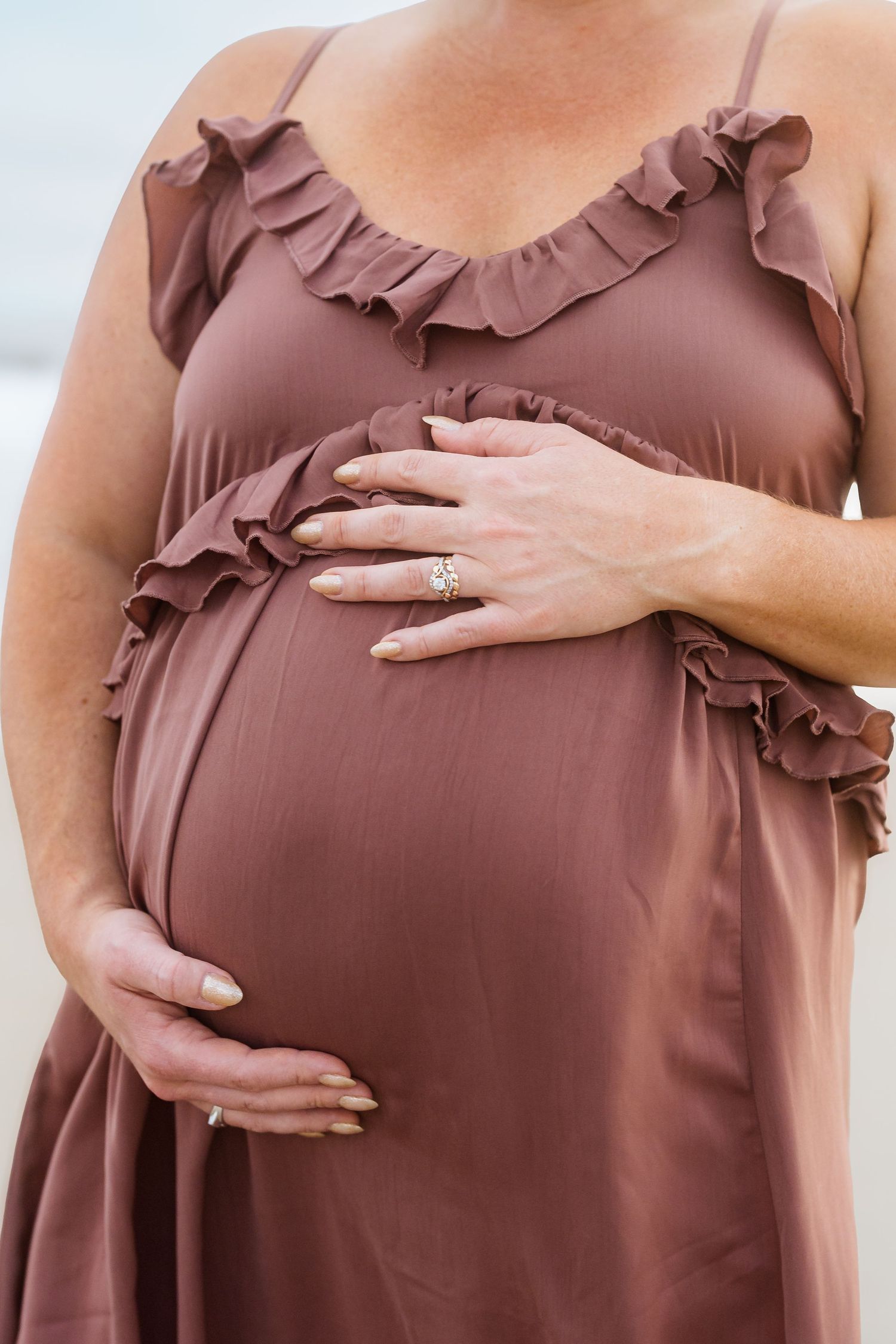Detail shot of hands tenderly cradling baby bump in ruffled mauve maternity dress.