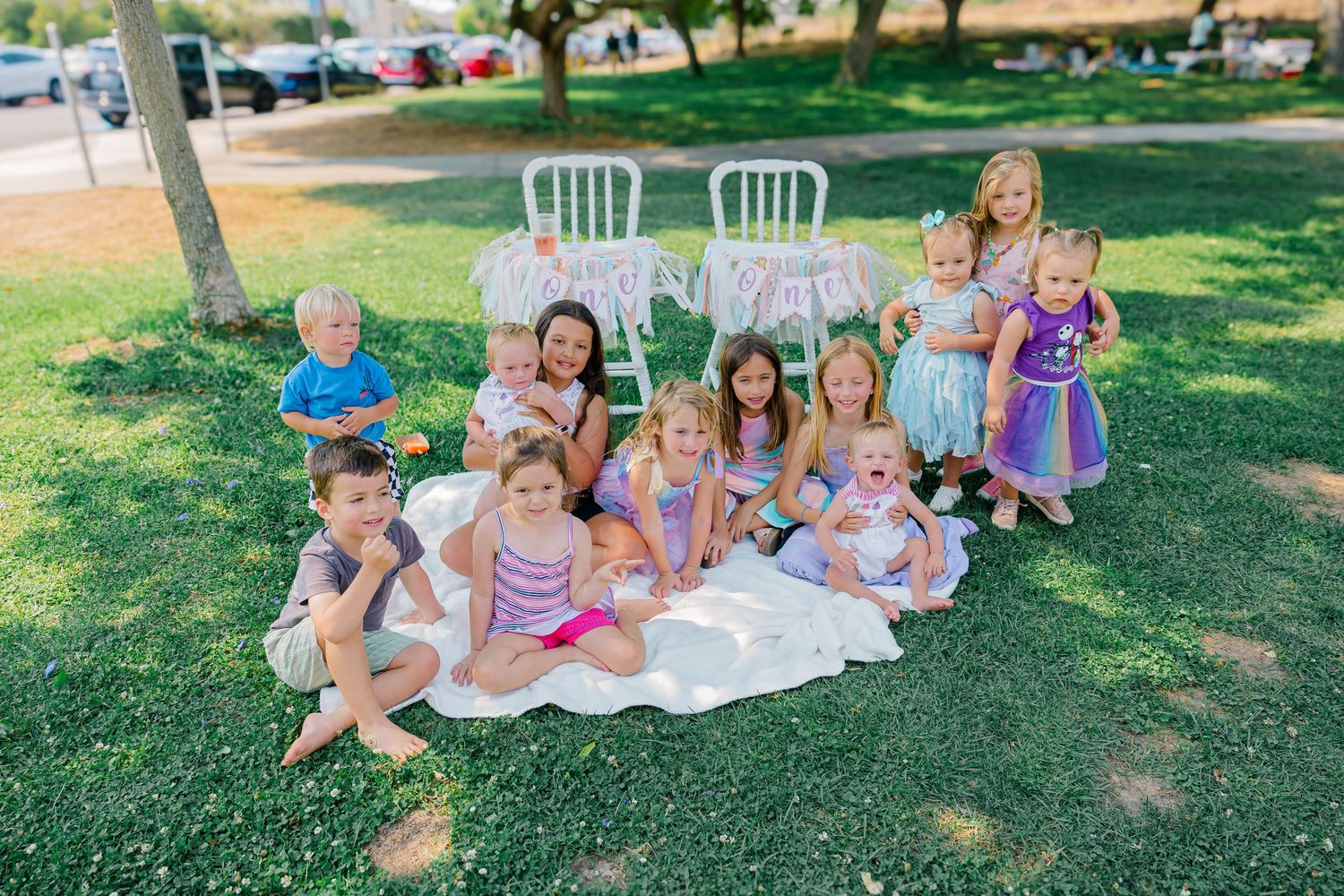 Children and adults gather on a blanket for an outdoor celebration in a grassy park setting.