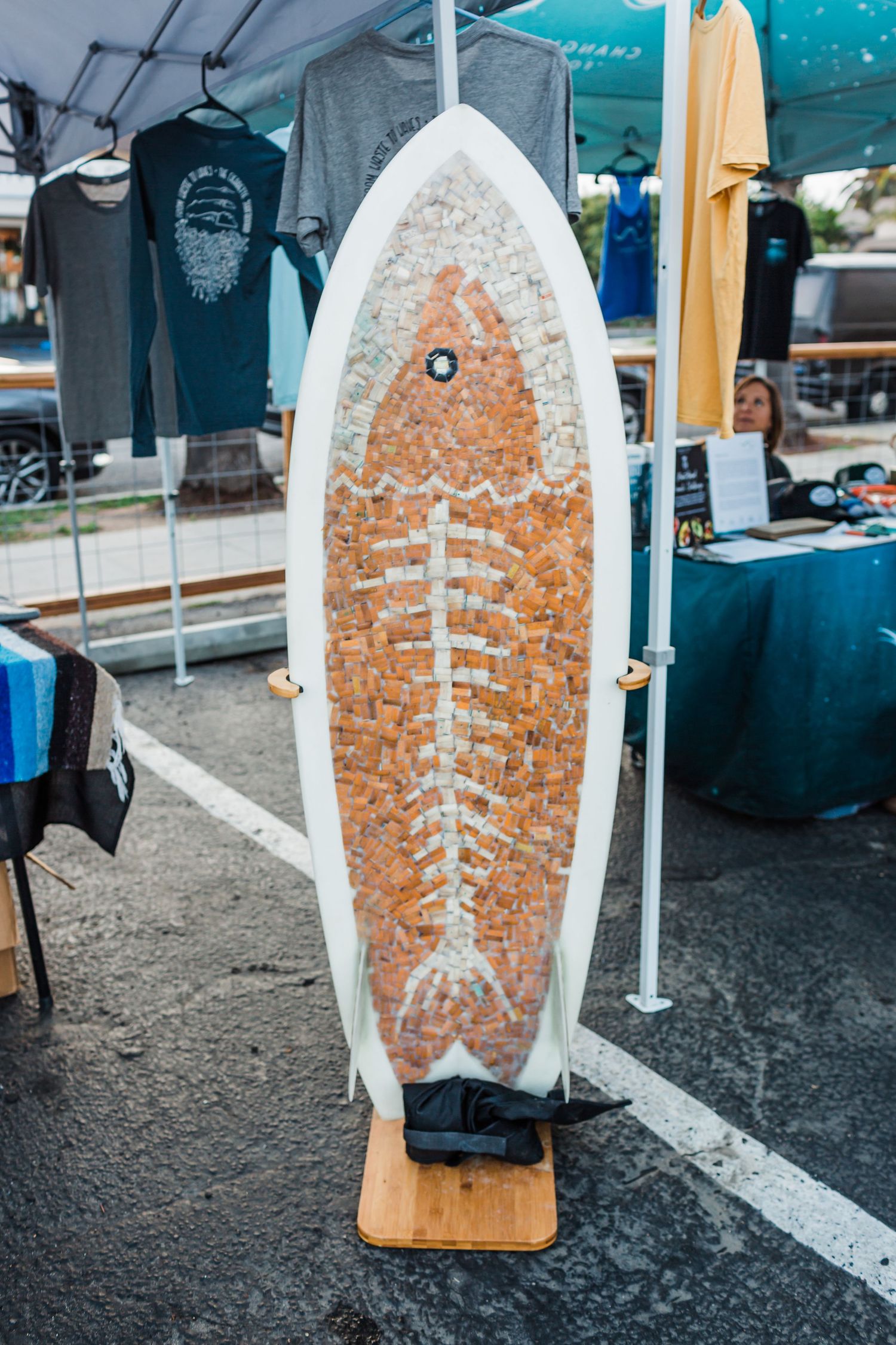 A rustic brown rusty surfboard with a painted cross design leans against a white table at a beach market.