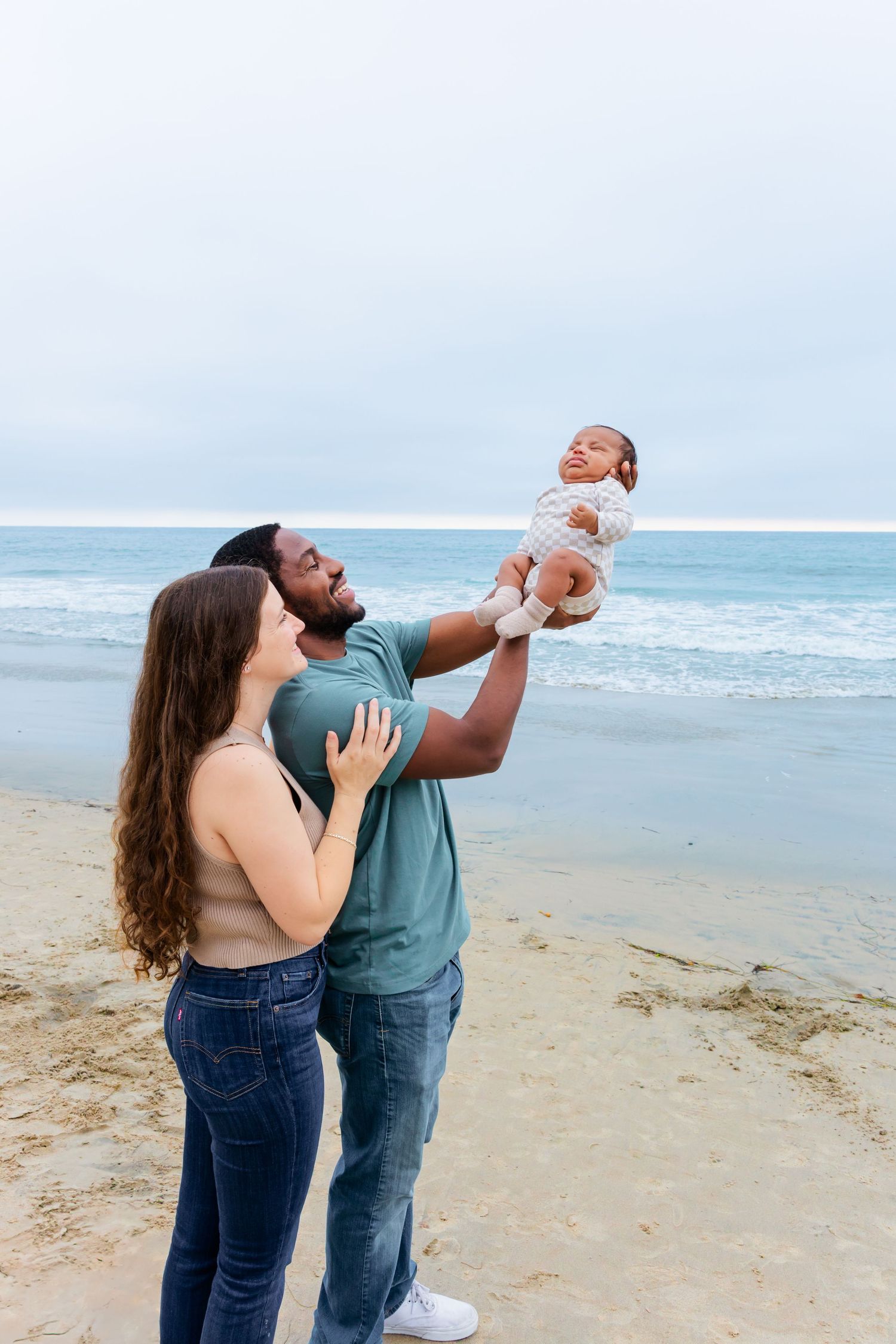 Parents playfully lifting their smiling baby in the air at the beach.