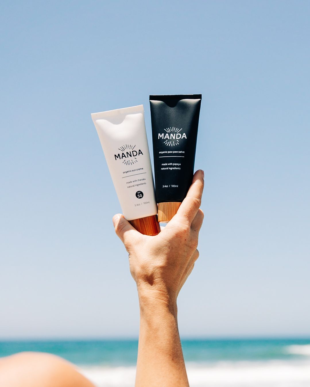 Two Manda sunscreen tubes held up against blue ocean background.