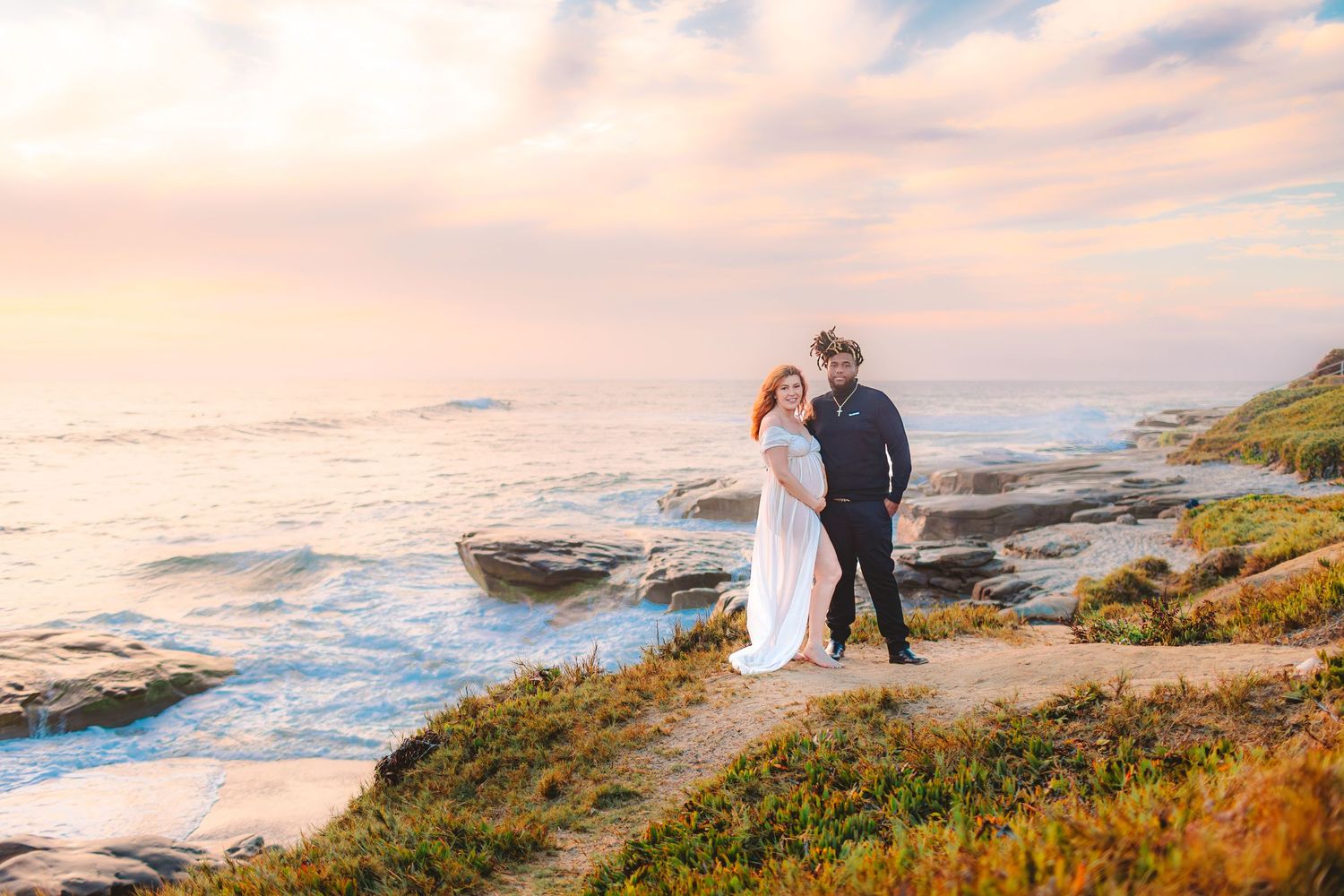 A couple walks along a scenic coastal cliff during golden hour as waves crash against the rocky shore below.