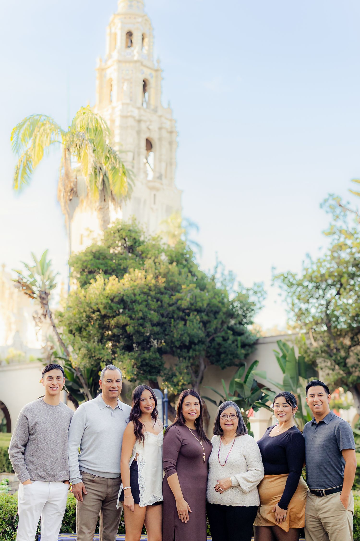 Extended family gathering posed together outdoors with white Spanish-style building and palm trees in background.