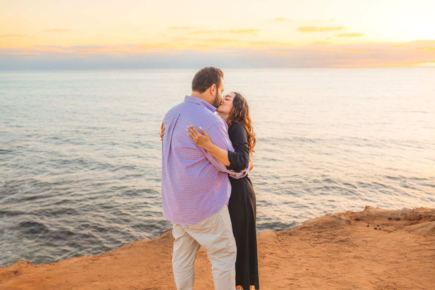 Silhouettes of a couple against an orange sunset sky on a dramatic oceanside cliff.