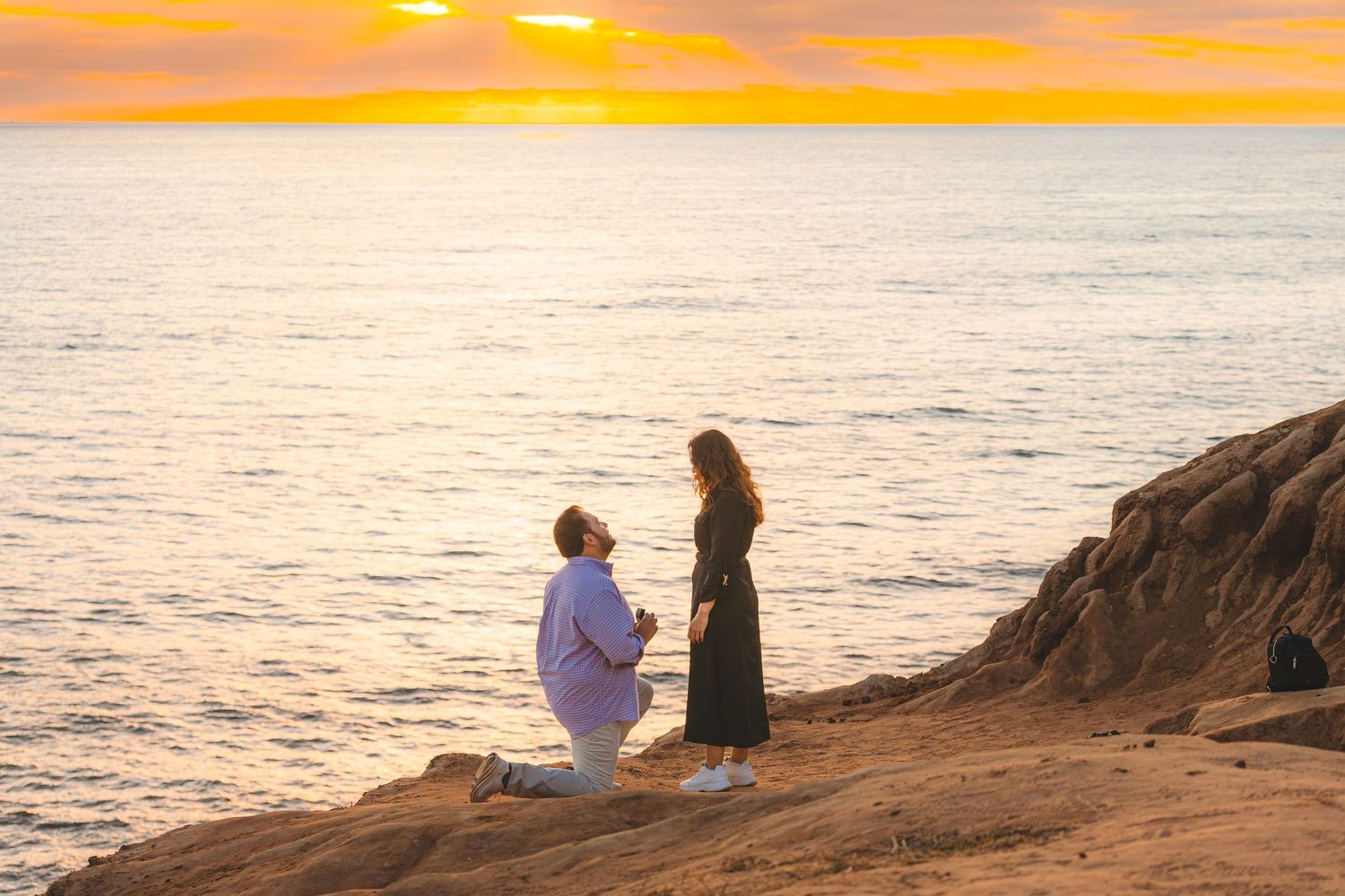 Silhouetted figures sit on rocky coastal cliffs at sunset as golden light illuminates the ocean horizon.