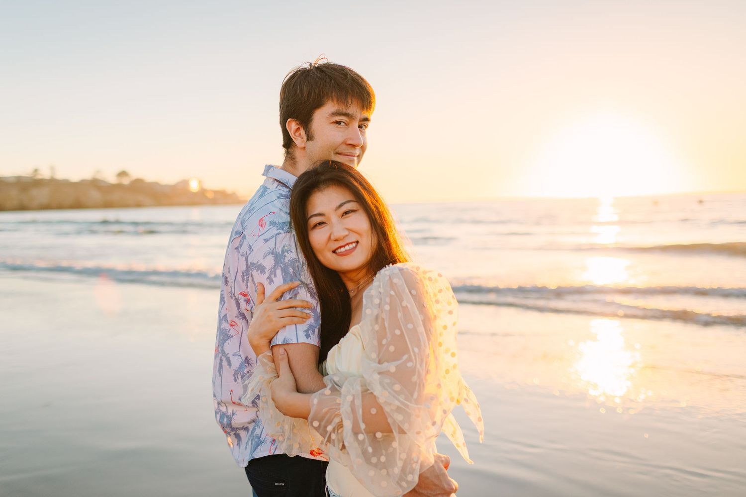 A couple embraces on a sunny beach at sunset with ocean waves in the background.