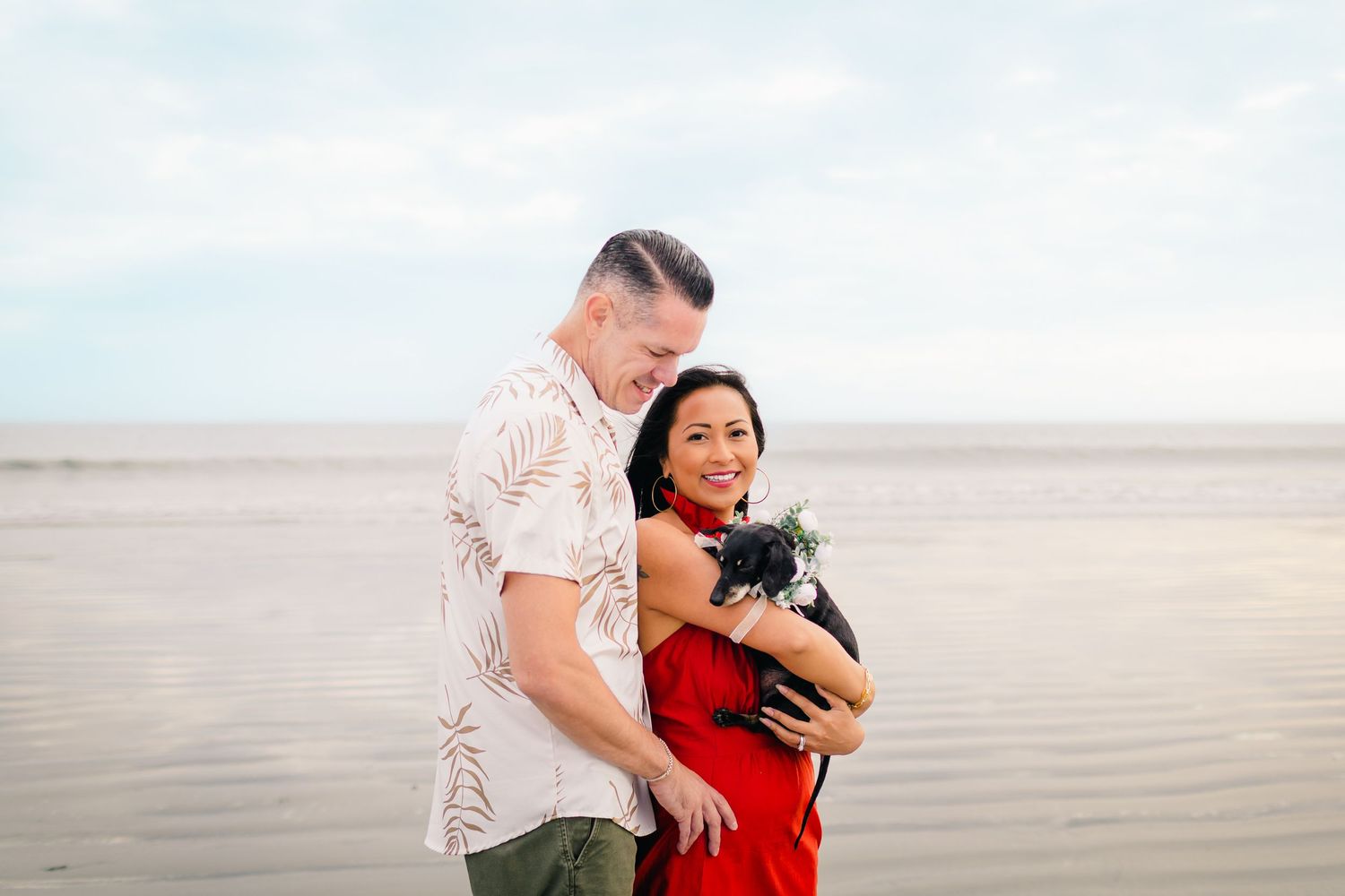 A couple in casual attire holds their small black dog while standing together on a misty beach.