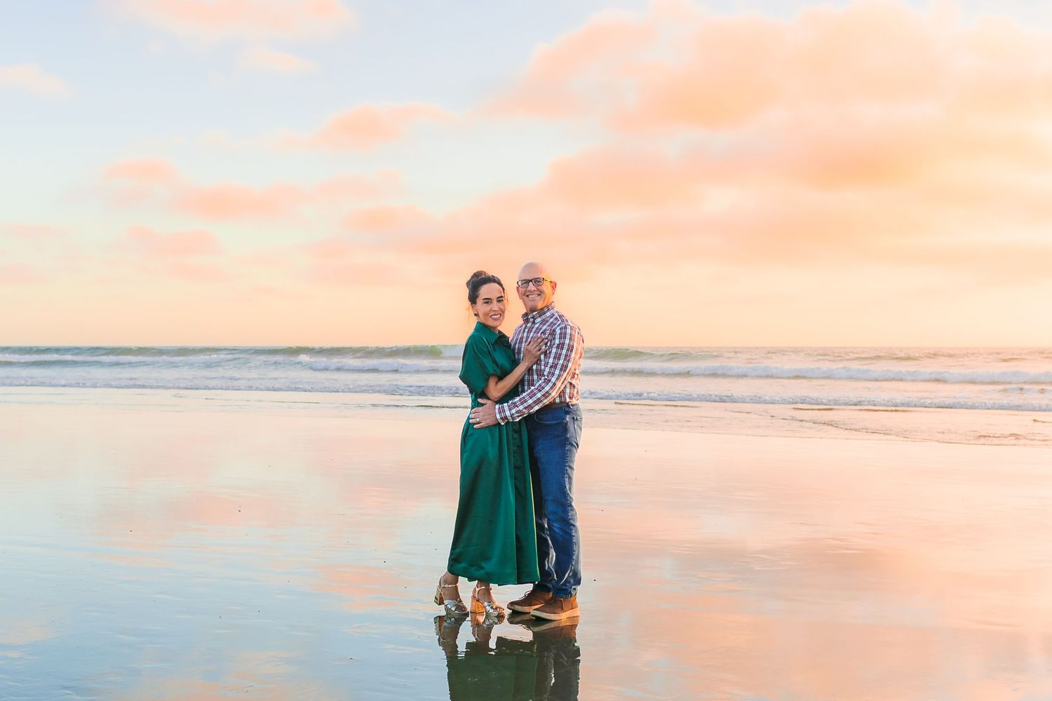 Couple embracing on sandy beach during golden hour with ocean waves and pastel sky in background.