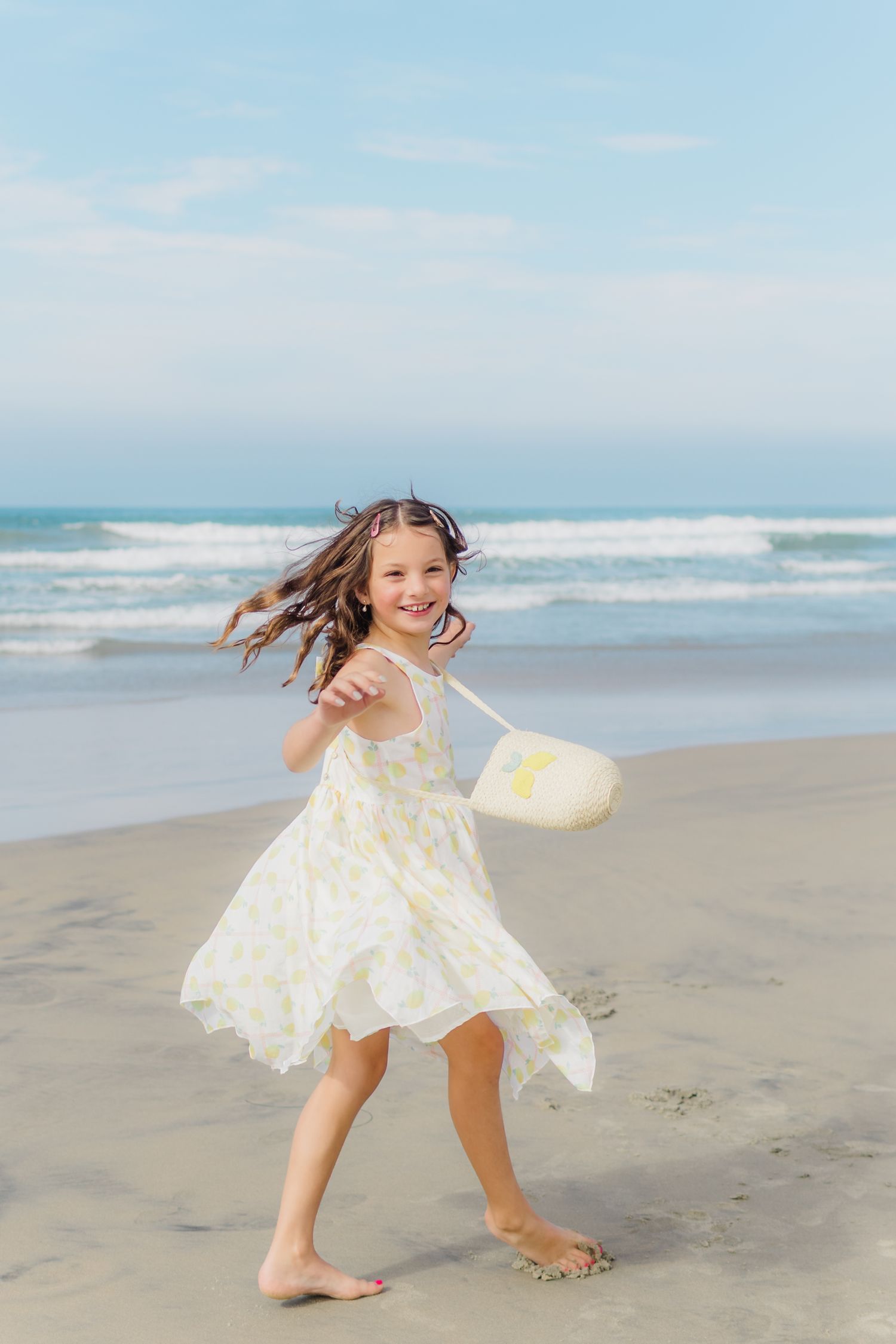 Young child in white dress twirling and dancing joyfully on a sunny beach with ocean waves in background.