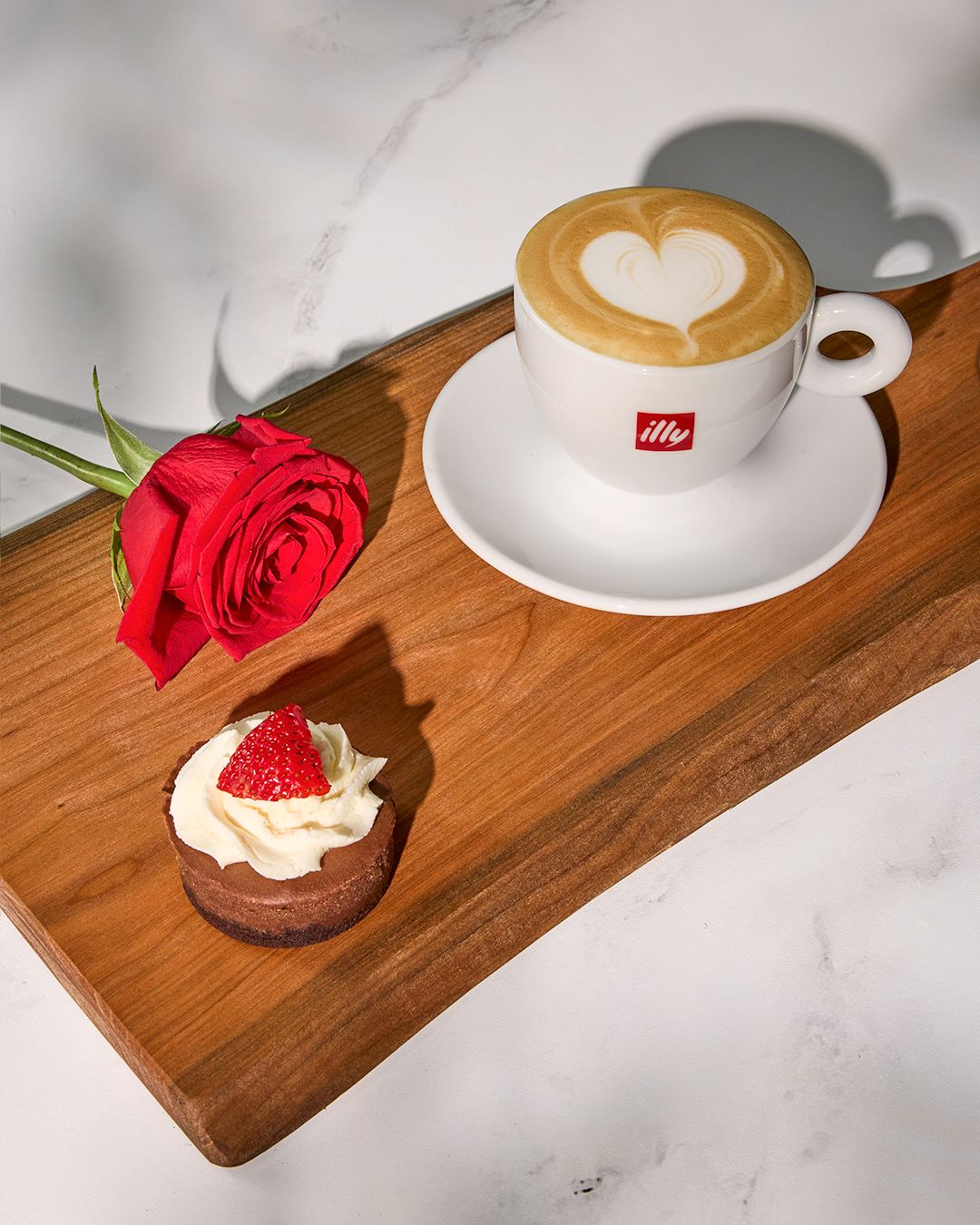 Latte with heart art, red rose, and chocolate dessert arranged on wooden serving board against marble background.