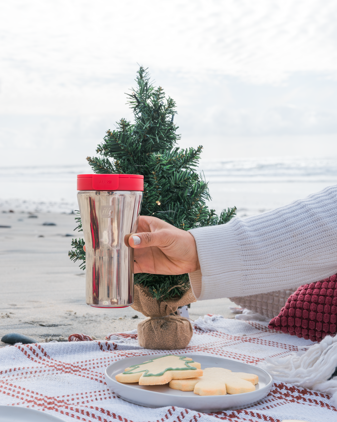 Holiday scene with silver travel mug and cookies by miniature Christmas tree on beach blanket.