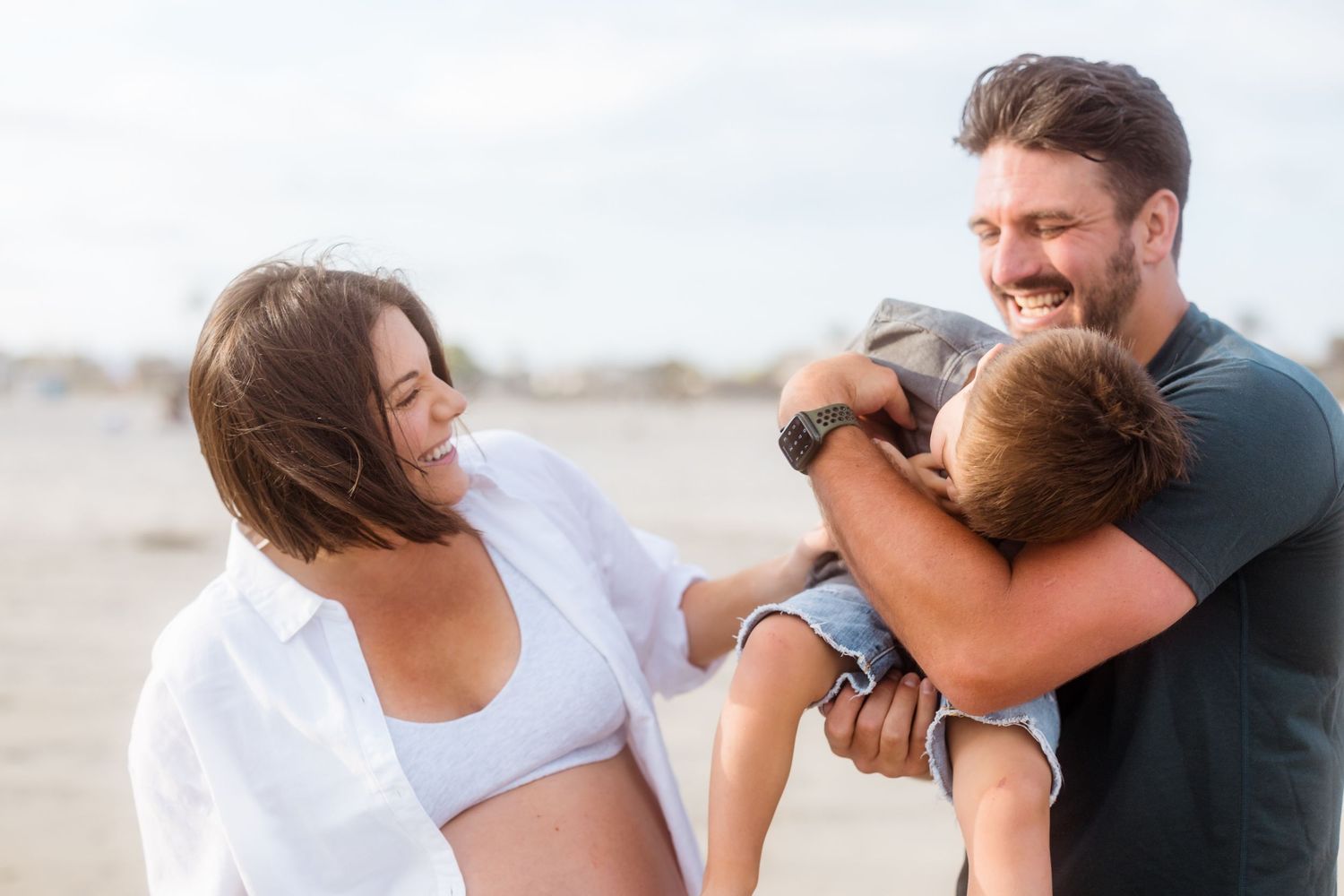 Happy family moment captured on beach with toddler and expecting parents.