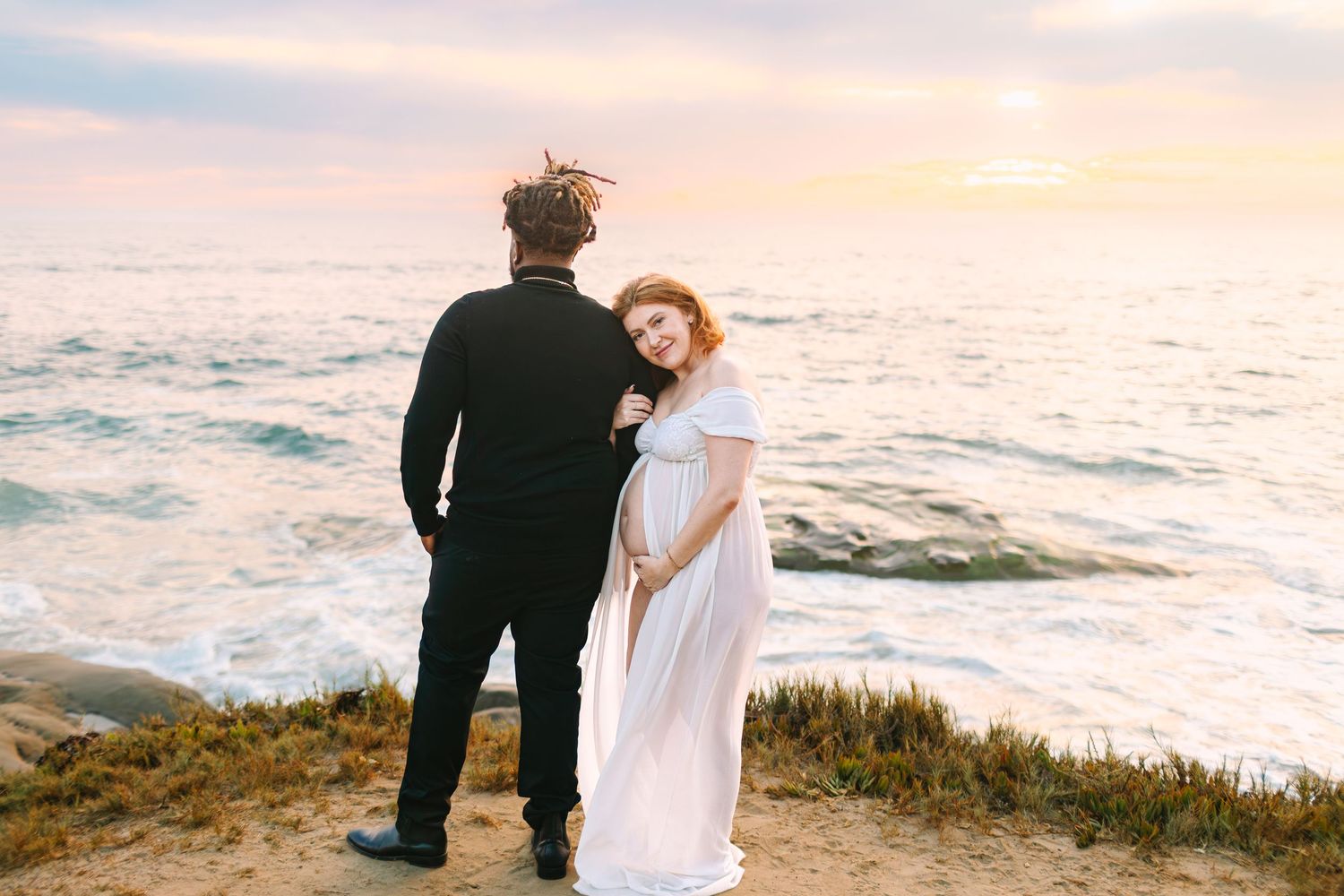 Romantic coastal wedding photoshoot featuring couple in formal attire embracing by the ocean at sunset.
