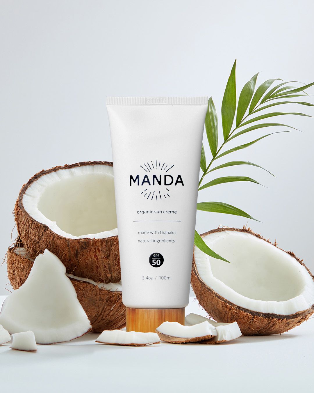 White Manda skincare tube displayed with fresh coconut pieces and palm leaf on blue background.