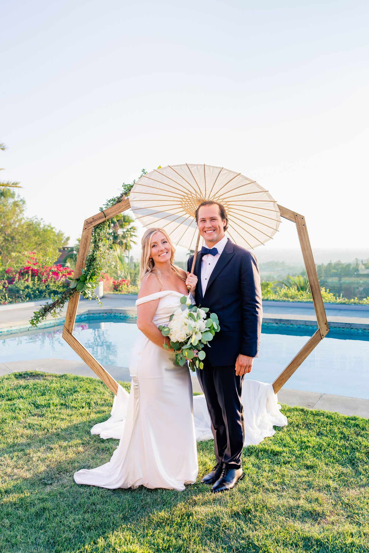 Wedding couple poses with decorative parasols on a grassy hillside overlooking water.