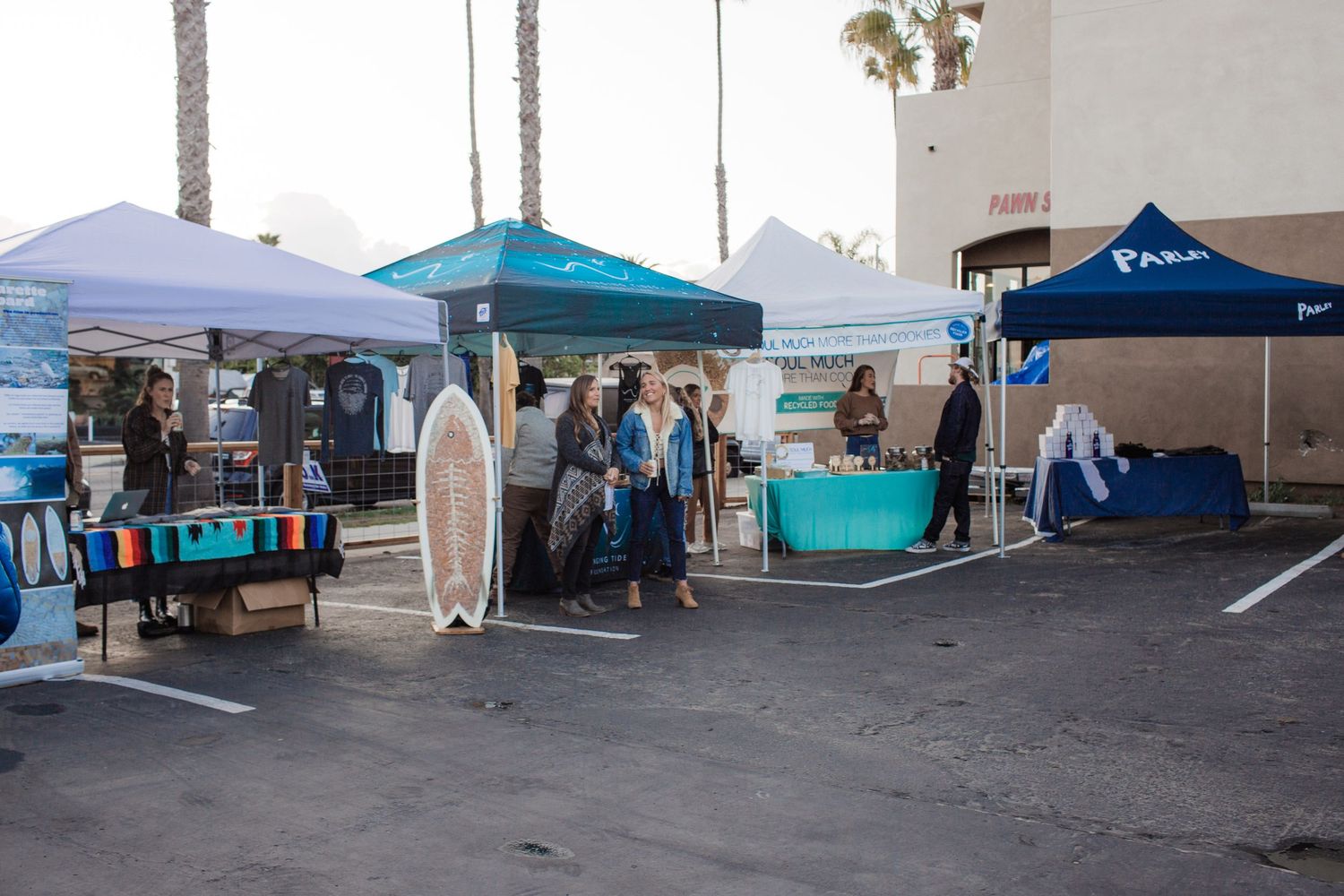 Outdoor market stalls with pop-up tents, surfboards and vendors set up in a parking lot during daytime.
