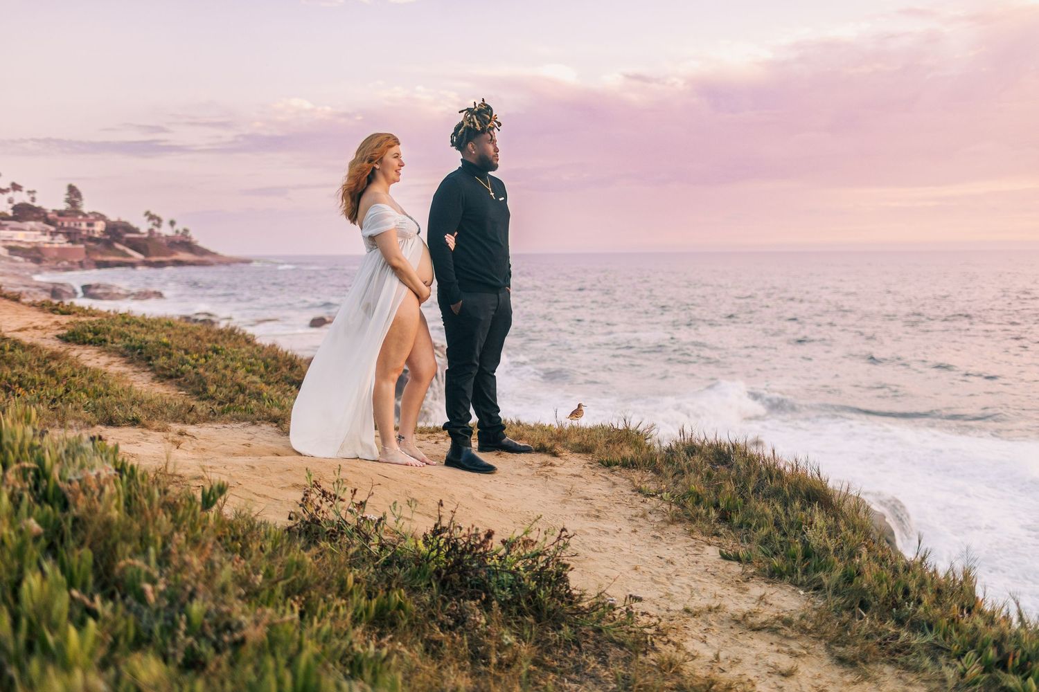 A couple in formal wear stands on a coastal path during sunset with the ocean and purple sky in the background.