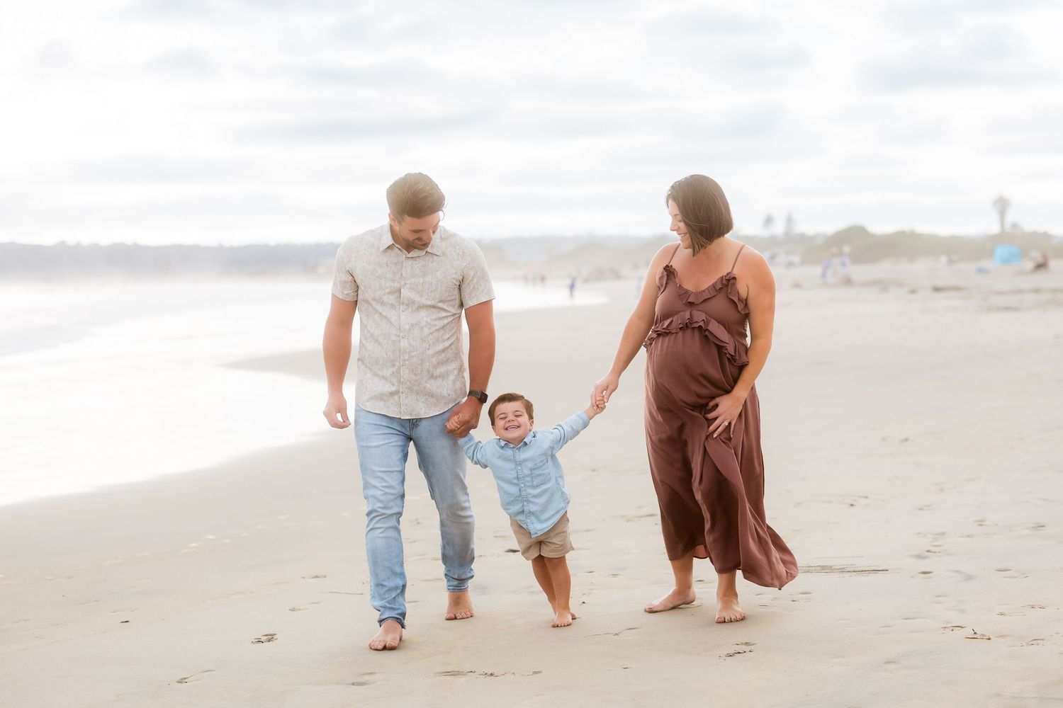 A family helps their toddler take steps on the beach during an overcast day.