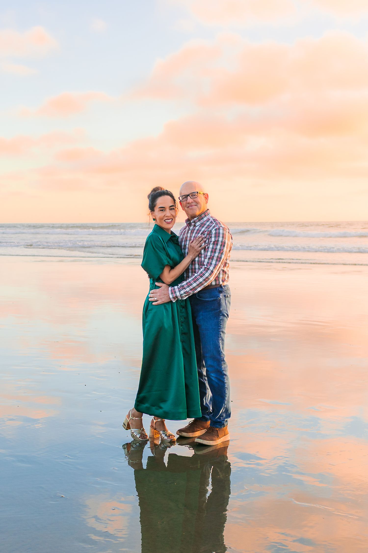 Couple embraces on beach at sunset with soft pink sky reflecting in wet sand and ocean waves.