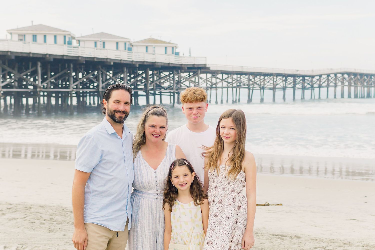 A group of people walking and smiling together on a sandy beach with a wooden pier in the background.