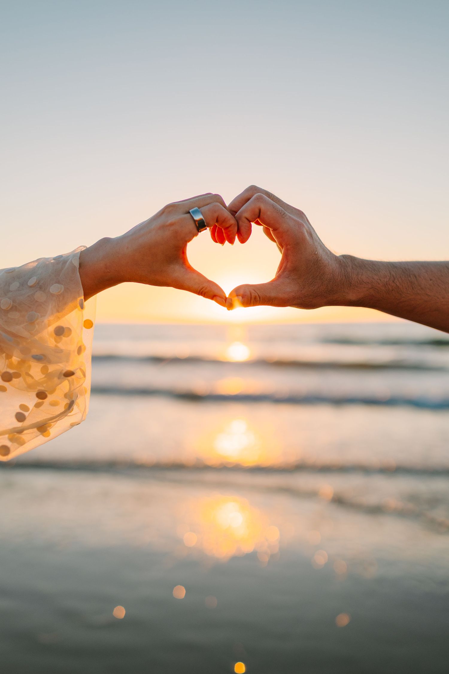 Hands forming heart shape silhouetted against golden sunset over ocean waves.