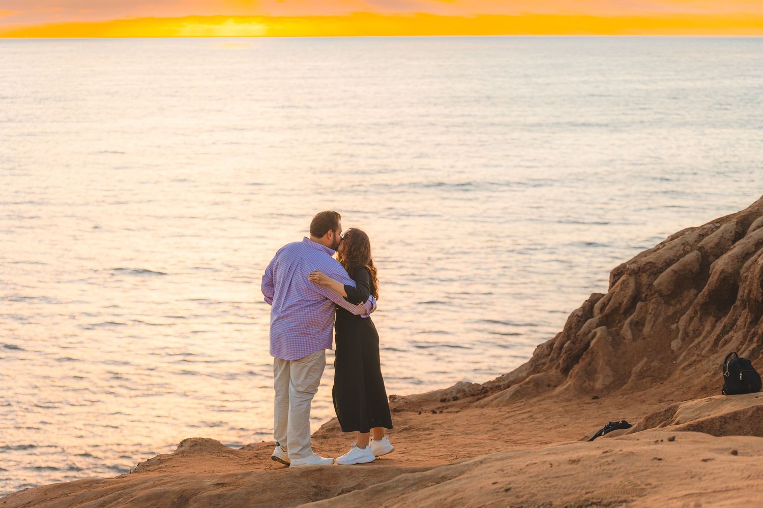 Two people embrace while watching the golden sunset from a rocky oceanside cliff.