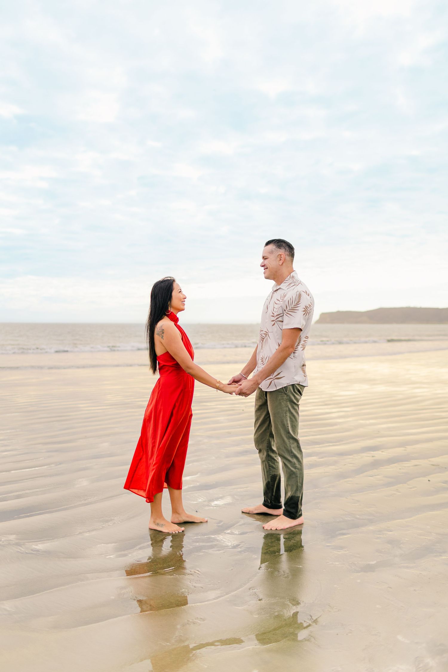 A couple stands together in shallow ocean water, with the person in red dress holding hands with their partner.