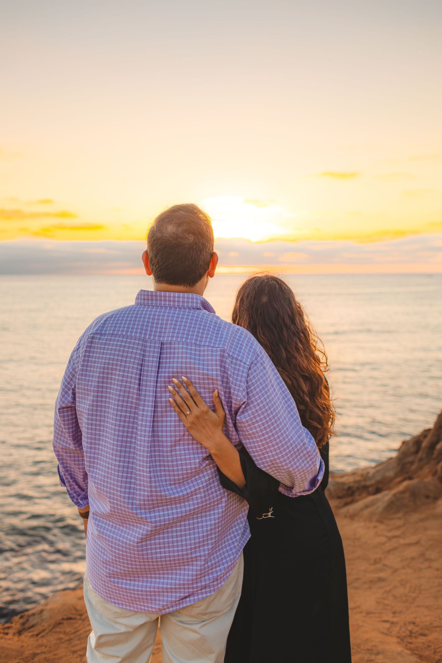 A couple watches the sunset together while sitting on a coastal cliff overlooking the ocean.