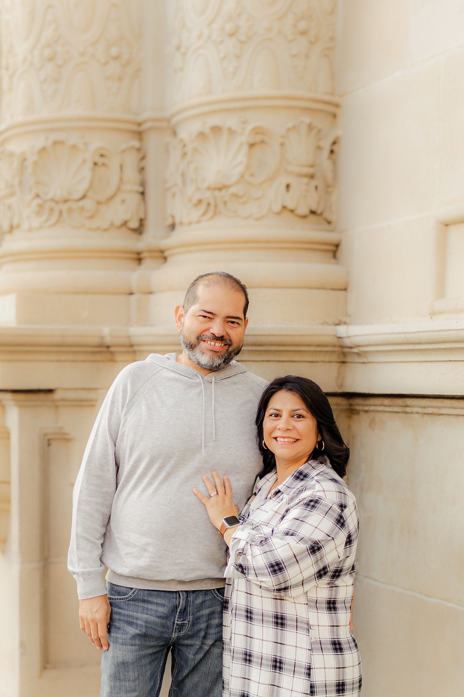 A couple shares joyful moments while posing against ornate white stone columns in a casual photoshoot.