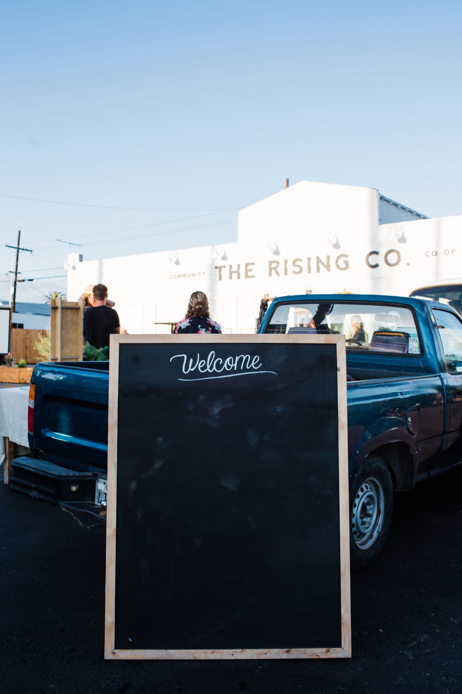 Old classic car parked in front of rustic welcome sign at The Rising Co establishment at dusk.