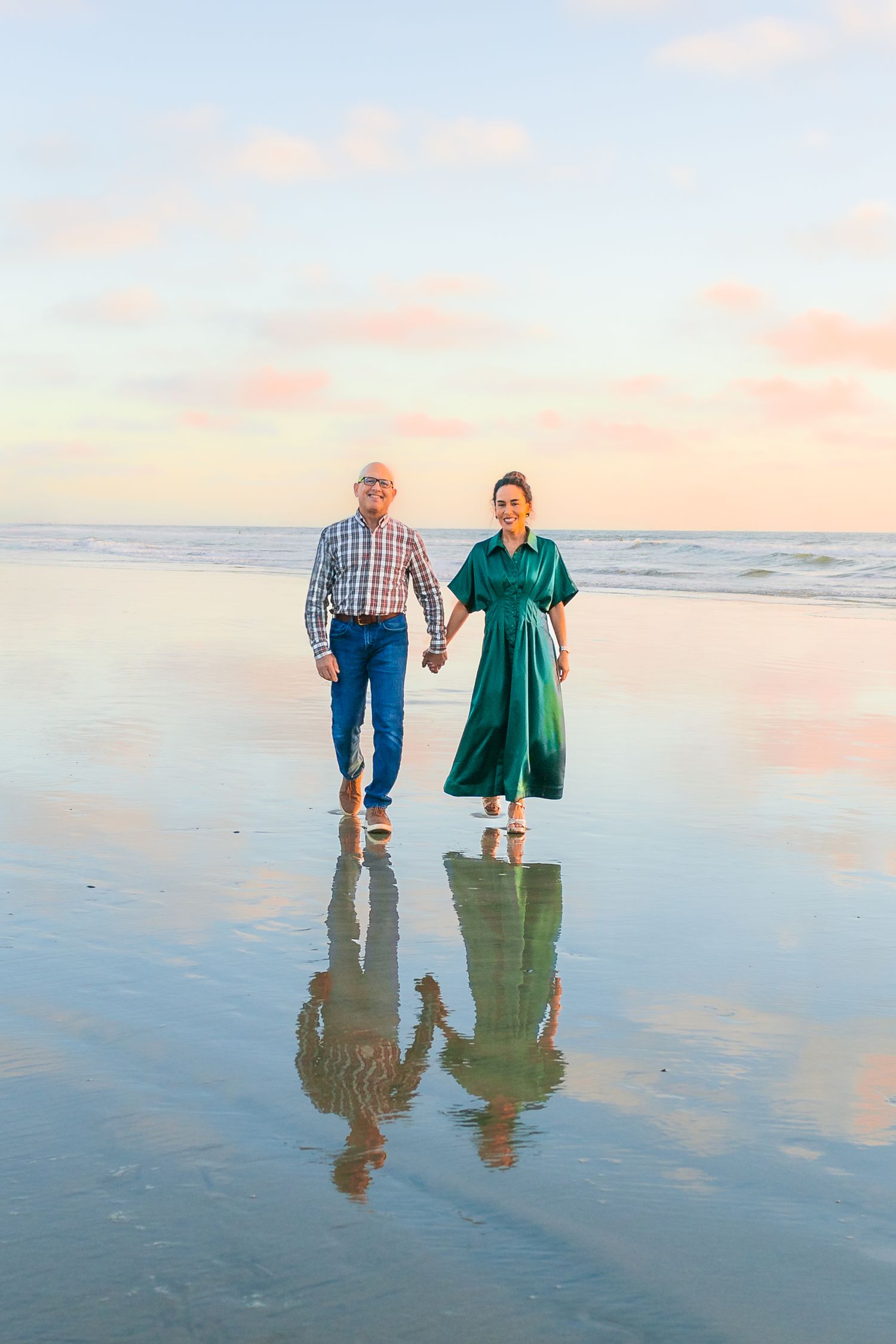 Two people walk hand in hand on a reflective beach at sunset with their mirror images showing in the wet sand.