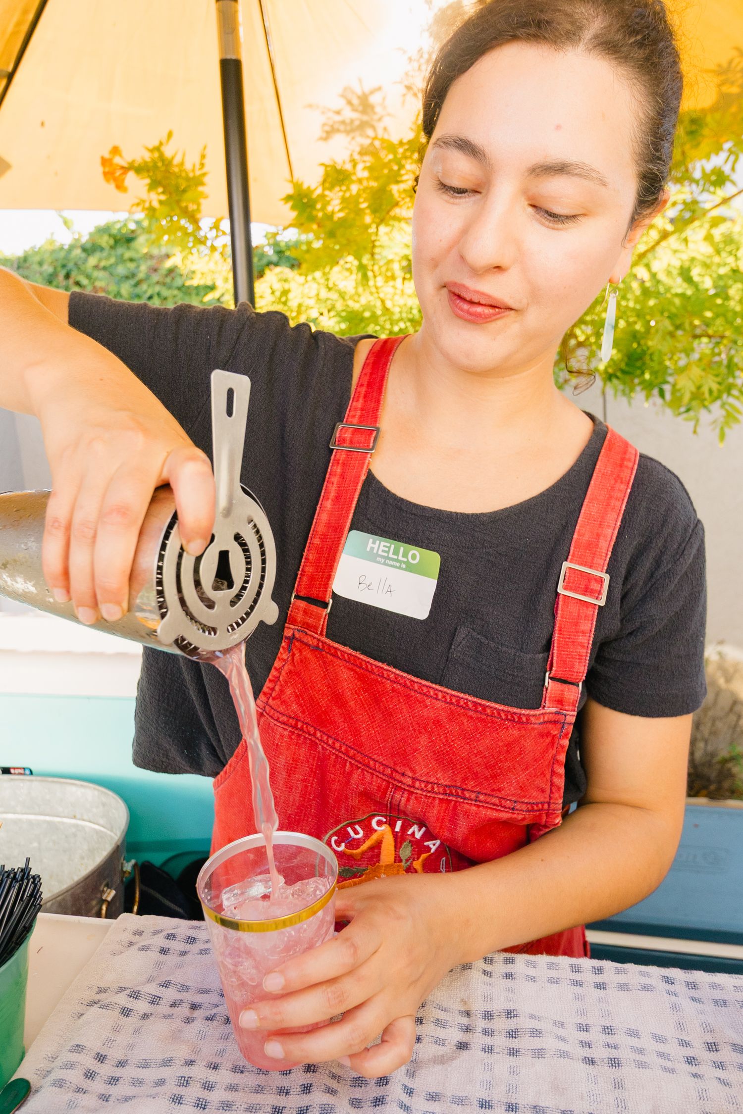 Person in red overalls pours a colorful drink into a clear glass on a checkered tablecloth.