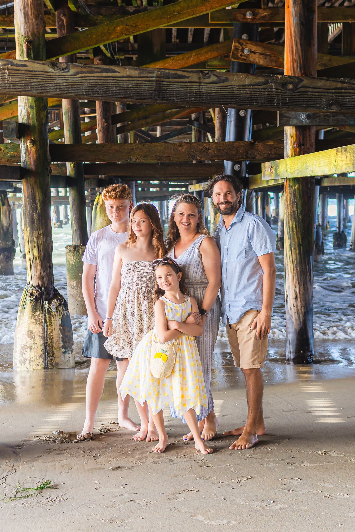 A family poses together under a wooden pier on a sandy beach during a casual beach photo session.