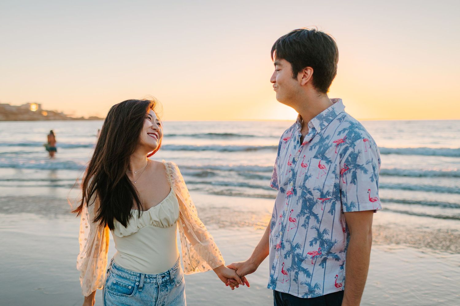 Romantic couple holding hands and walking along beach at sunset wearing matching floral prints and denim.