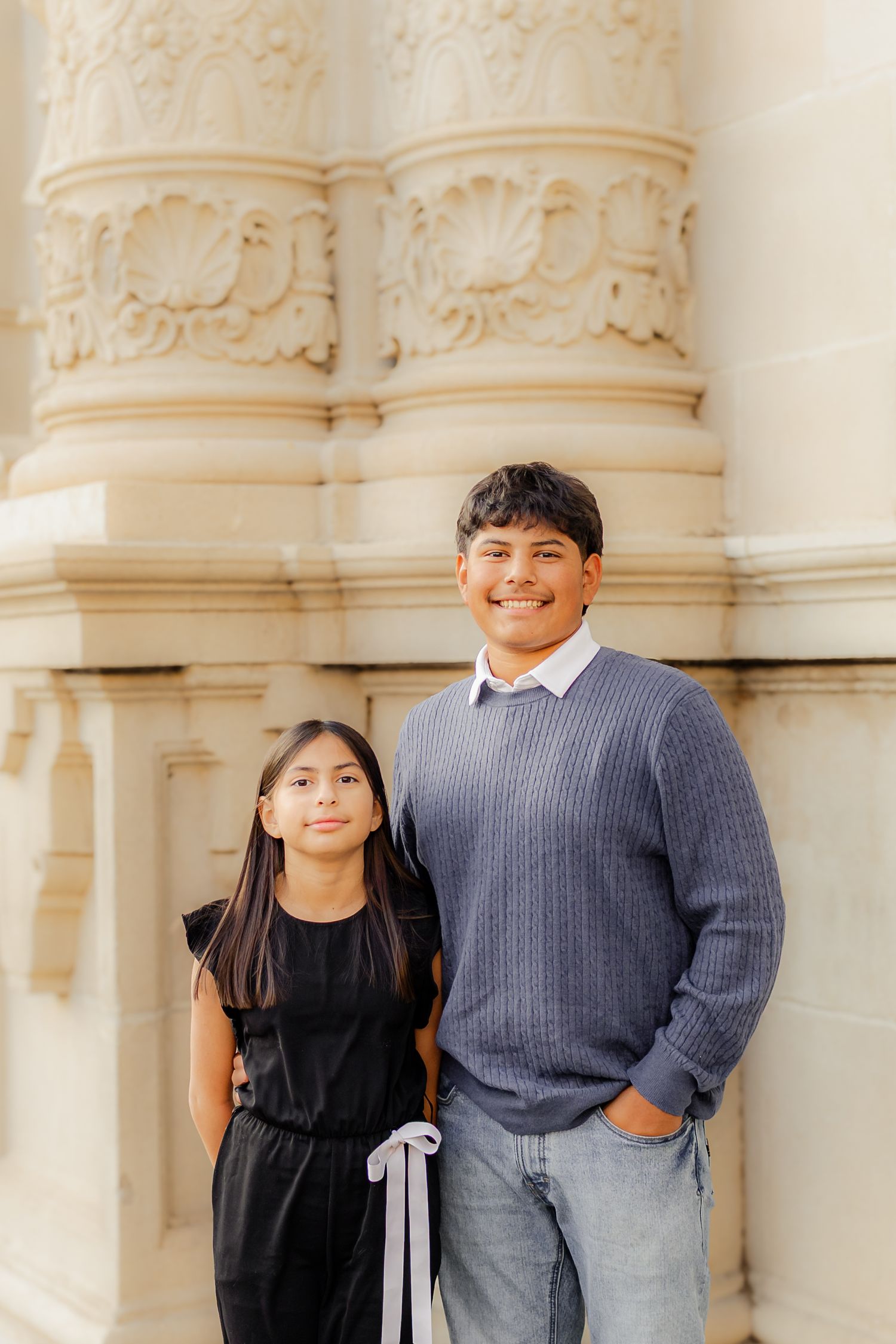 Two children in formal attire pose against ornate white stone columns at an elegant architectural setting.