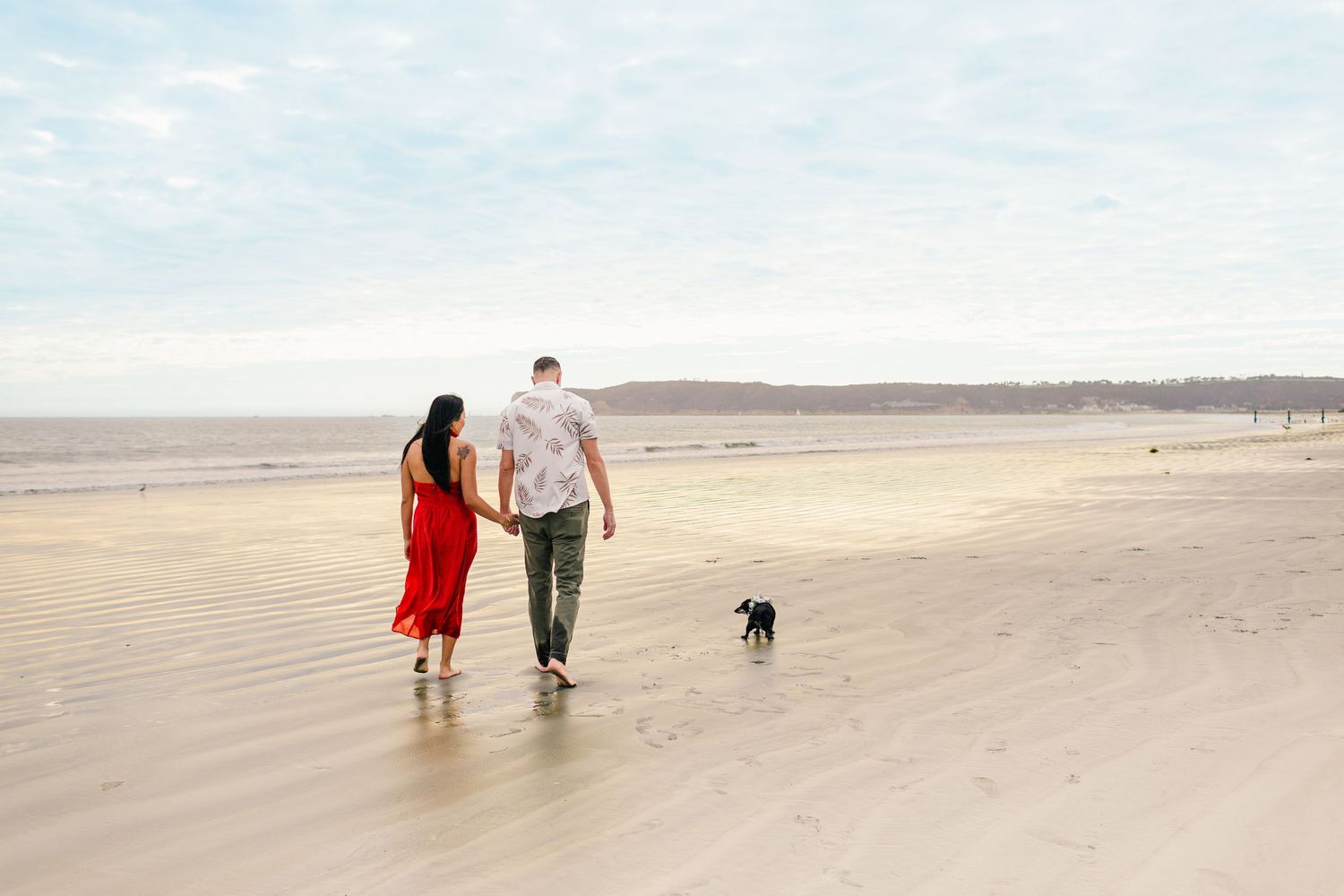 Silhouettes of people walking together on a wide sandy beach against a bright cloudy sky.