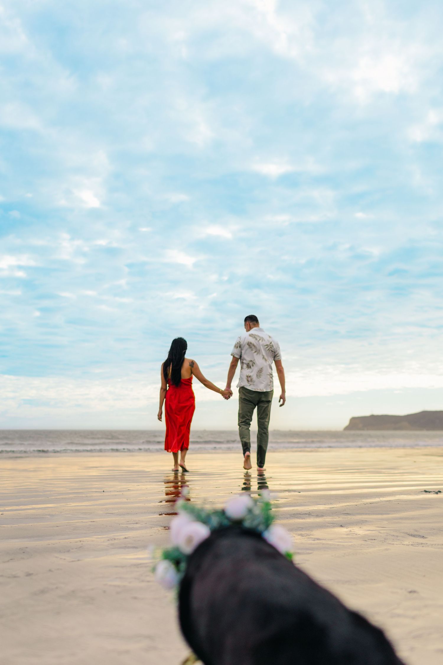 Two people stroll hand in hand on a serene beach with dramatic cloudy skies above.